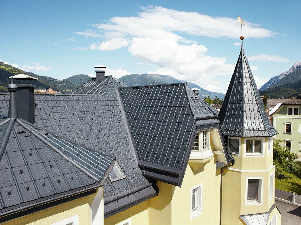 Buildings with many details, towers and angled roof surfaces; the roof is covered with PREFA’s roof tile in anthracite. The PREFA half-round gutter and the PREFA on-roof gutter in anthracite are used for roof drainage.