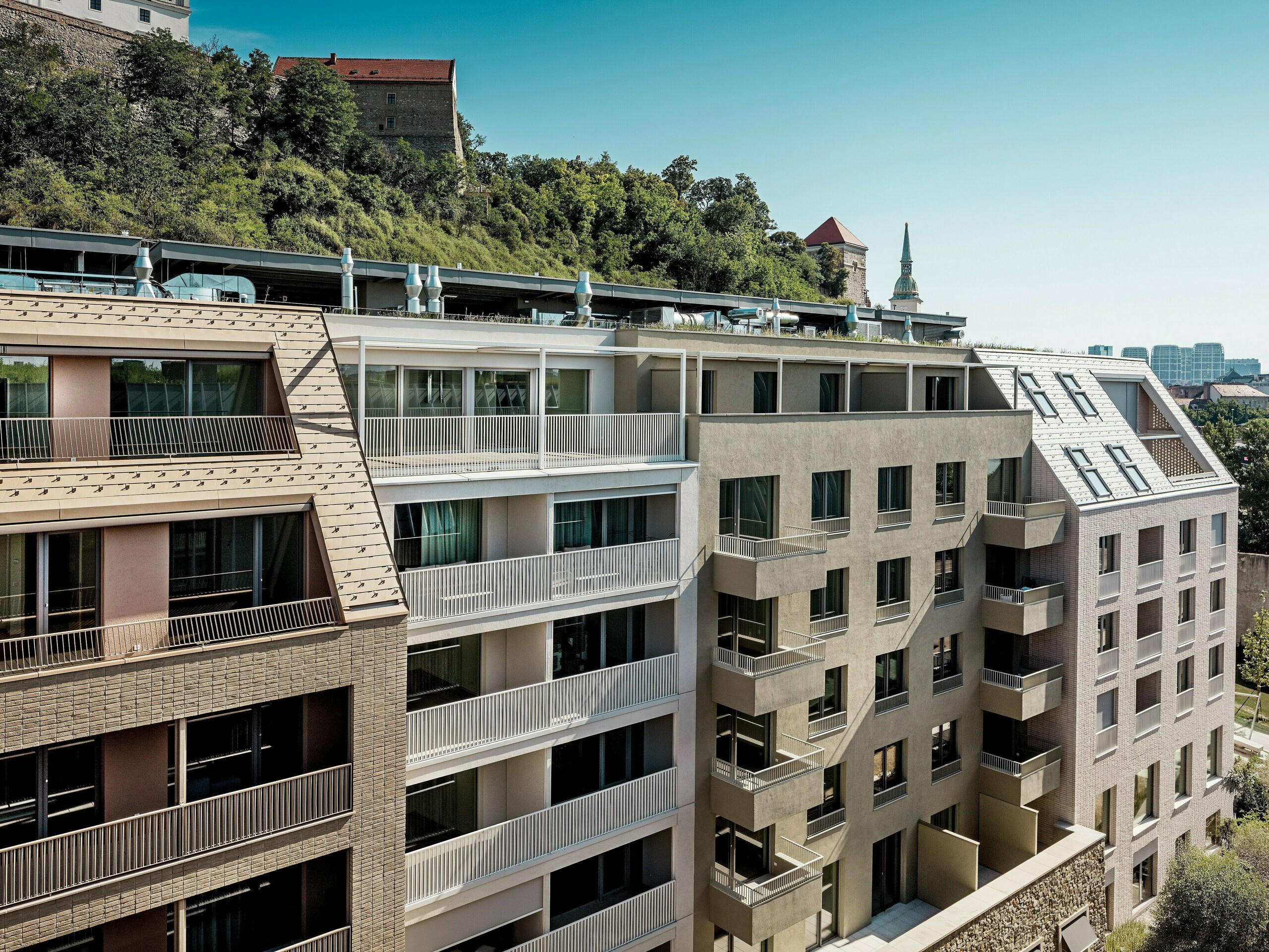 Urbanes Wohnensemble im Quartier Vydrica in Bratislava mit eindrucksvollem Zusammenspiel aus moderner Architektur und historischer Kulisse nahe der Burg: Die steilen, präzise gestalteten Dächer mit PREFA Aluminium Dachschindeln in Sandbraun und Prefaweiß bilden mit ihren klaren Linien, Gauben und großzügigen Dachterrassen die visuellen Highlights. Die hochwertigen Aluminiumlösungen ermöglichen komplexe Dachformen, überzeugen durch Langlebigkeit, Witterungsbeständigkeit und geringen Wartungsaufwand und schaffen eine nachhaltige, ästhetisch anspruchsvolle Gebäudehülle im urbanen Kontext.