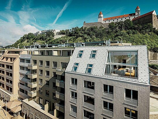 Moderne Dachlandschaft im Stadtquartier Vydrica in Bratislava mit Blick zur Burganlage: Steil geneigte Dachflächen mit großzügigen Fensteröffnungen und klaren Linien werden von PREFA Aluminium Dachschindeln in Prefaweiß und Sandbraun elegant in Szene gesetzt. Die präzise Verarbeitung und die harmonische Verbindung von Dach und Fassade schaffen einen starken visuellen Eyecatcher über den Dächern der Stadt. PREFA Aluminium überzeugt durch Leichtigkeit, hohe Witterungsbeständigkeit und langlebige Qualität – ideal für komplexe, architektonisch anspruchsvolle Wohnbauten.