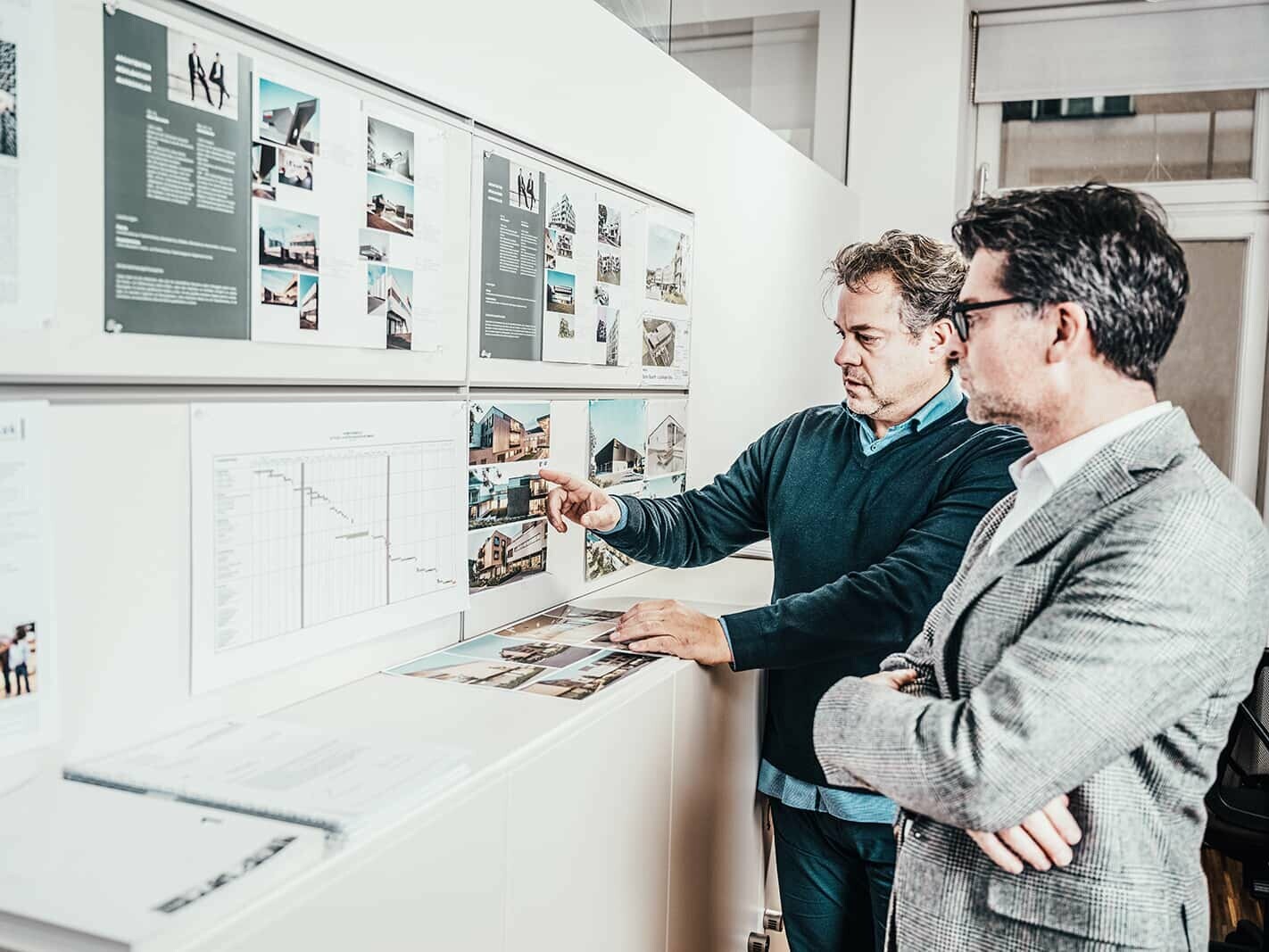 Picture of the architects Mühlbacher and Marschalek. They are looking on a pin board which is covered with pictures of houses.