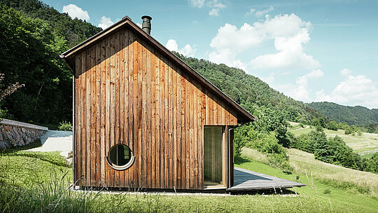 Minimalistisches Holzhaus mit runder Fensteröffnung und langlebigem PREFALZ Aluminiumdach in P.10 Braun, eingebettet in die hügelige Landschaft von Vinje. Die PREFA Dacheindeckung ist wartungsfrei, bietet optimalen Schutz vor Wind und Wetter und harmoniert farblich mit der natürlichen Holzfassade. Sie unterstreicht die klare, zeitlose Architektur des Gebäudes. Von der großzügigen Terrasse aus hat man einen traumhaften Blick auf die Hügellandschaft Sloweniens. Die Perspektive zeigt die Stirnseite des Einfamilienhauses mit dem asymmetrischen Satteldach und den unterschiedlich großen Dachflächen.