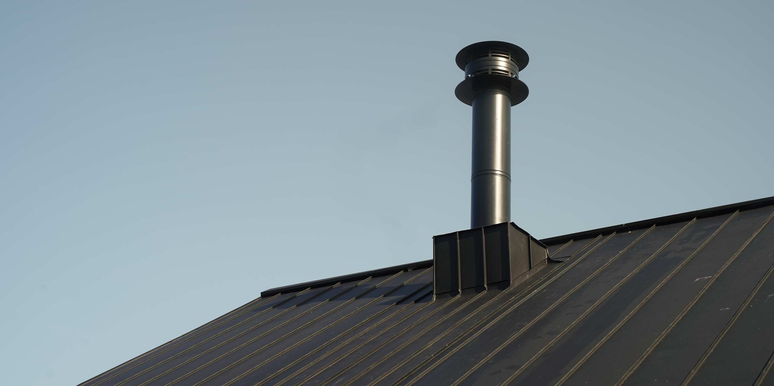 Close-up of a PREFALZ roof system in P.10 black on the "Golf it!" complex in Glasgow, Scotland, with a striking, modern chimney. The chimney rises elegantly above the precisely laid, dark standing seam sheets, which are known for their durability and robustness. This image underlines the combination of functionality and design that PREFA aluminium products offer.