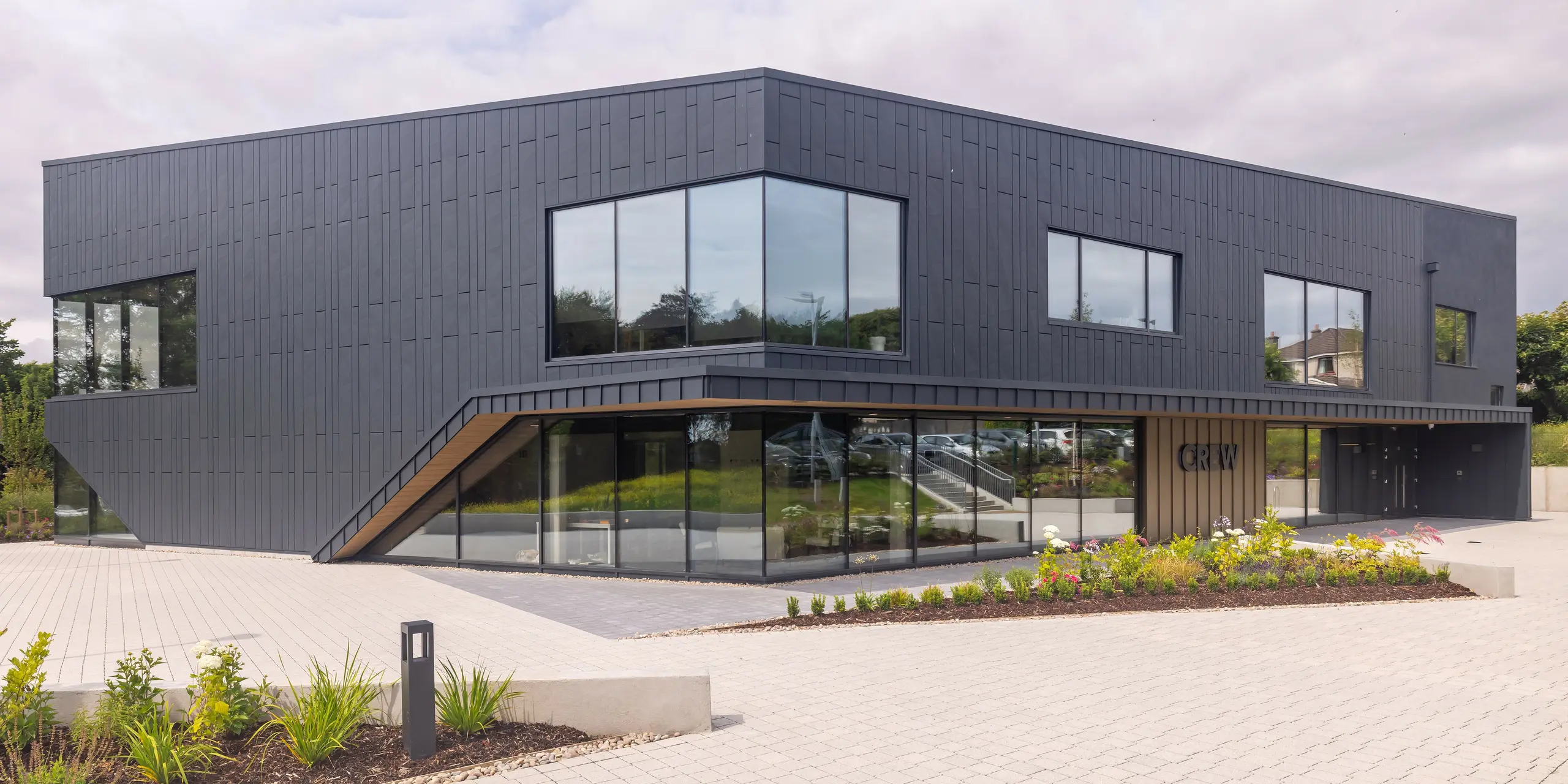 Front view of the CREW building in Galway, Ireland, with modern PREFA aluminium façade made of Siding.X and PREFALZ in P.10 anthracite. The angled lines and large window areas characterise the architecturally sophisticated design of the building. The anthracite-coloured aluminium cladding in combination gives the building an elegant and contemporary aesthetic, which is complemented by the neat landscaping.
