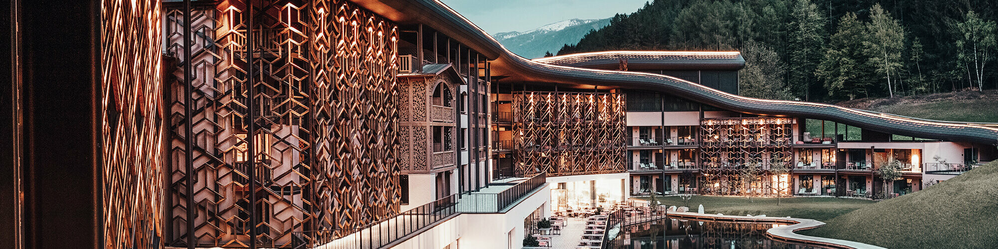 Evening shot, warm light illuminates the wooden façade and the aluminium roof, below the terrace with seating, in front of the lake, the green forest in the background