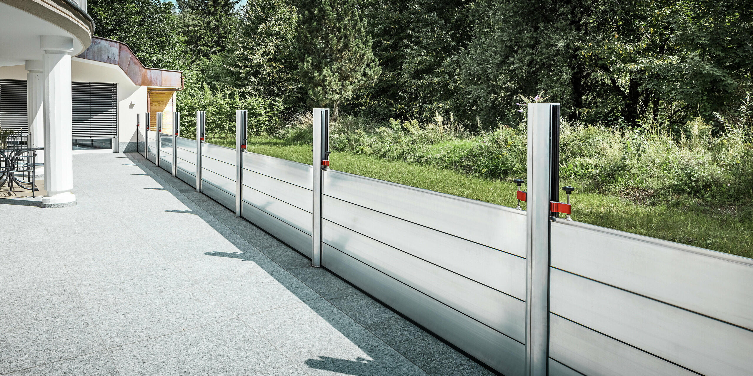 Seitenansicht der fertig montierten PREFA Hochwasserschutzwand entlang der Terrasse eines Wohnhauses in Tirol. Die robusten Dammbalken aus Aluminium sind zwischen vertikalen Stützen in Bodenhülsen befestigt und schützen das Grundstück vor möglichen Überflutungen aus dem angrenzenden Naturraum. Das System überzeugt durch präzise Verarbeitung, hohe Stabilität und unauffällige Integration in die moderne Architektur.