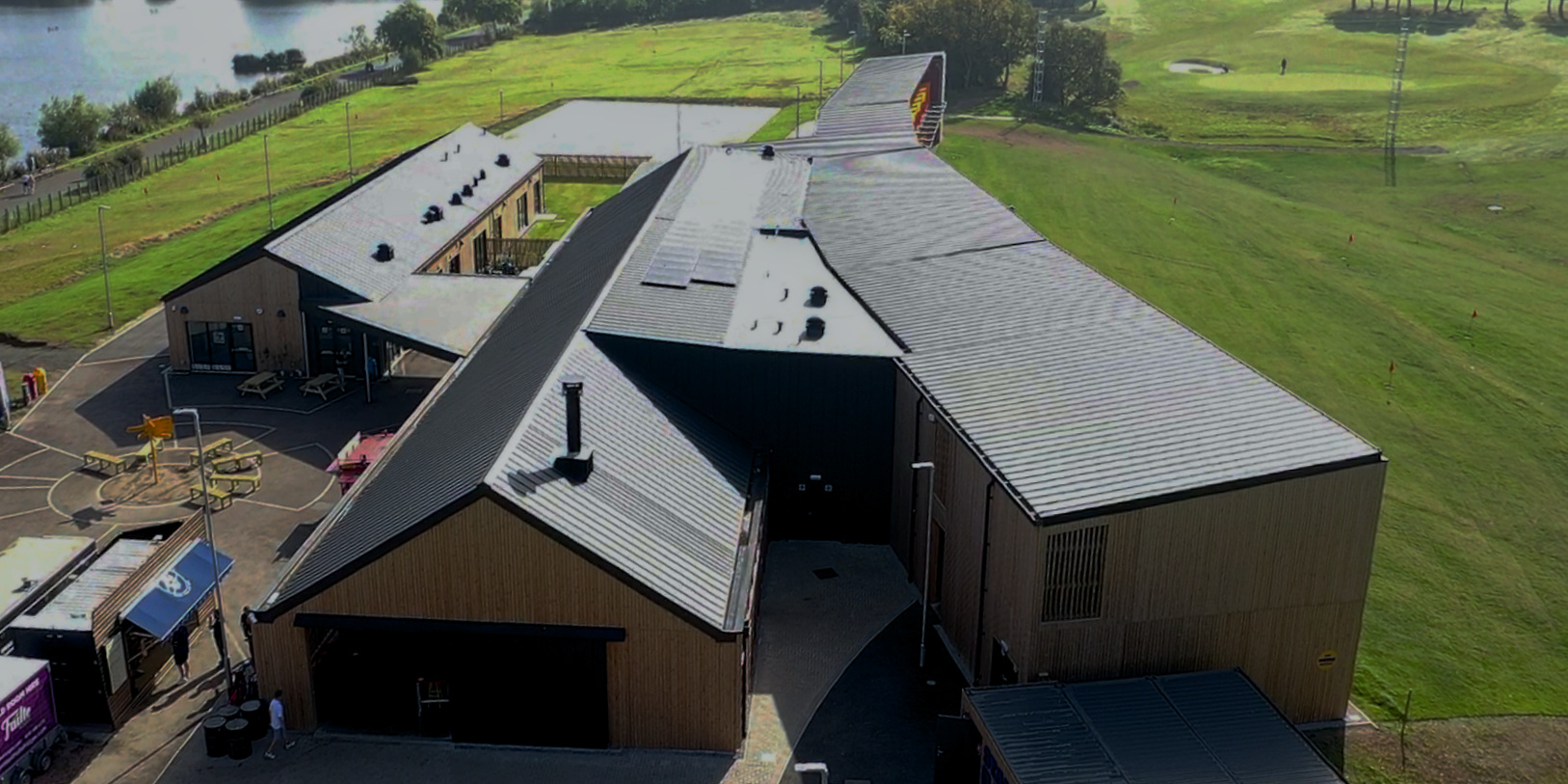The golf venue 'Golf it!' in Glasgow, embedded in a green landscape, has an impressive PREFALZ roof construction in P.10 black, which extends over several parts of the building. The roof, characterised by its clean lines and aesthetic design, reflects the modern and dynamic appearance of the golf course. The view from above also shows the neighbouring picturesque lake and the well-tended golf course, which emphasises the harmonious integration of the facility into the natural surroundings.