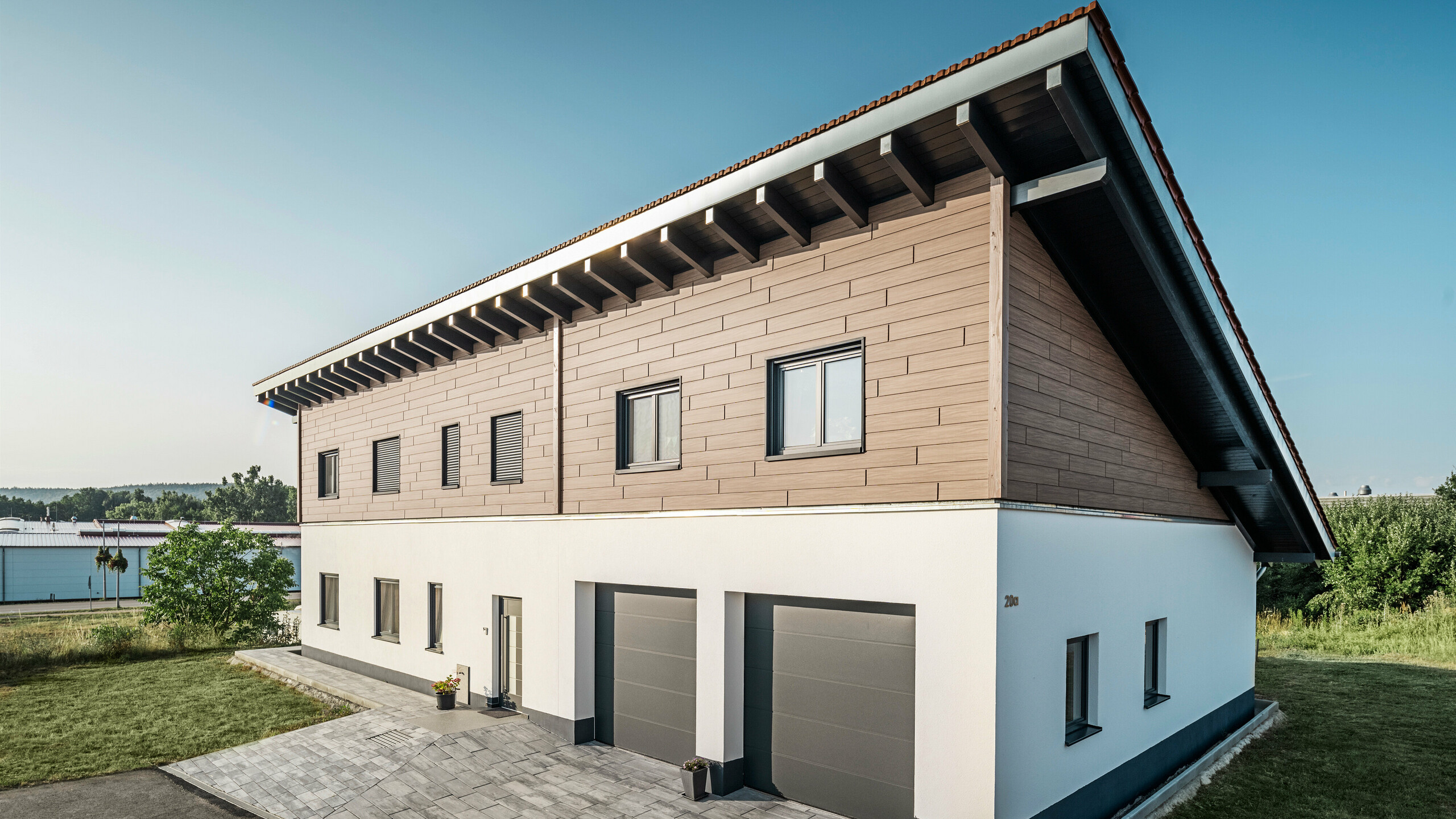 View from the side of a detached house with a monopitch roof in Bruck. The upper storey is clad with PREFA sidings in Walnut Brown. Two garage doors in anthracite can be seen on the right-hand side of the picture. The entrance door is located in the centre of the building. The plinth is in a plain white colour. 