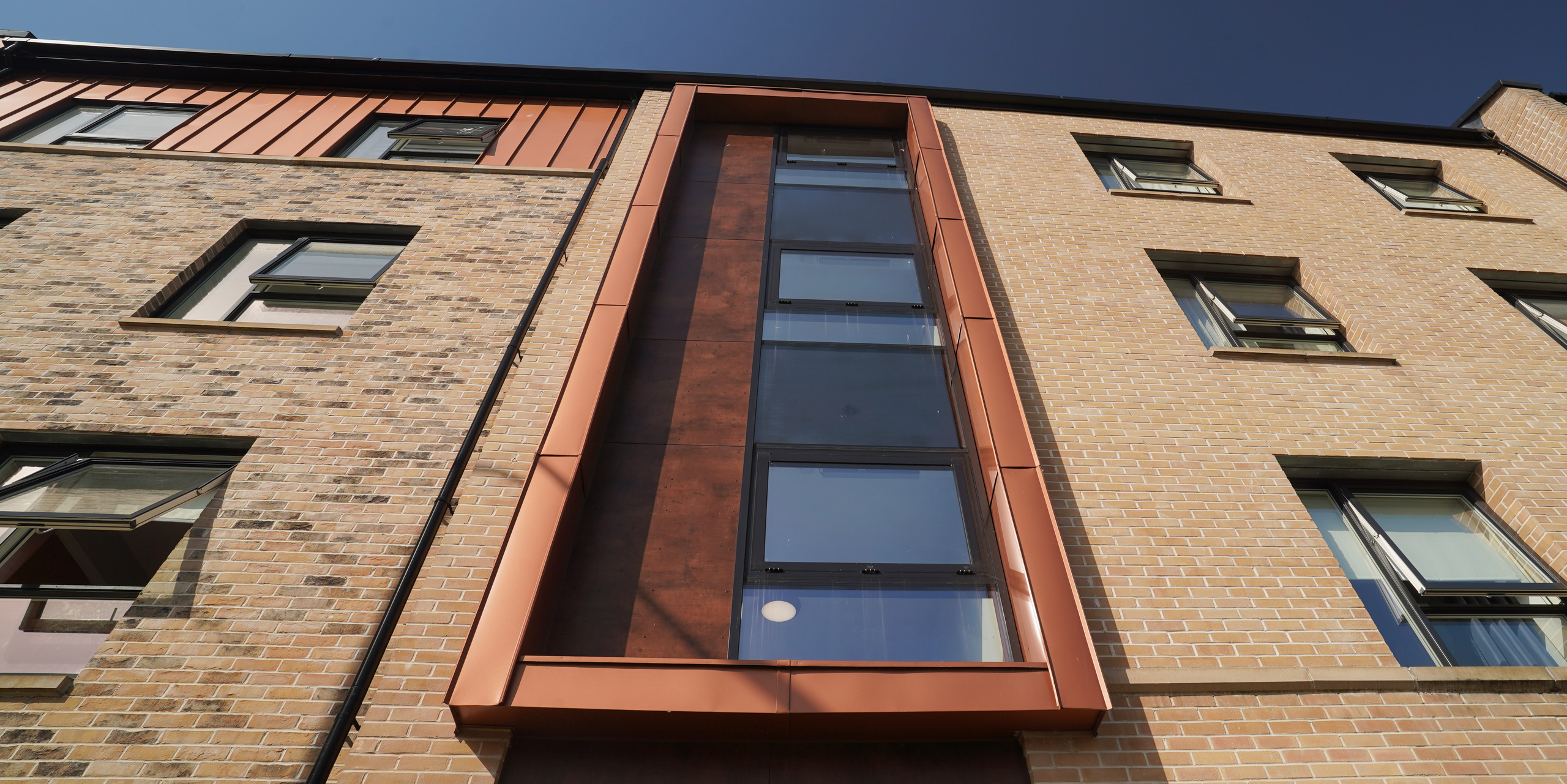 Detailed view of the façade of a residential building on Nethan Street in Glasgow, characterised by the cladding with FALZONAL in new copper. The copper-coloured aluminium cladding around the window sections sets modern accents against the classic brick façade and shows the interplay of innovative materials and traditional design. The high-quality aluminium product from PREFA underlines the aesthetics of the building and at the same time ensures its durability and weather resistance.