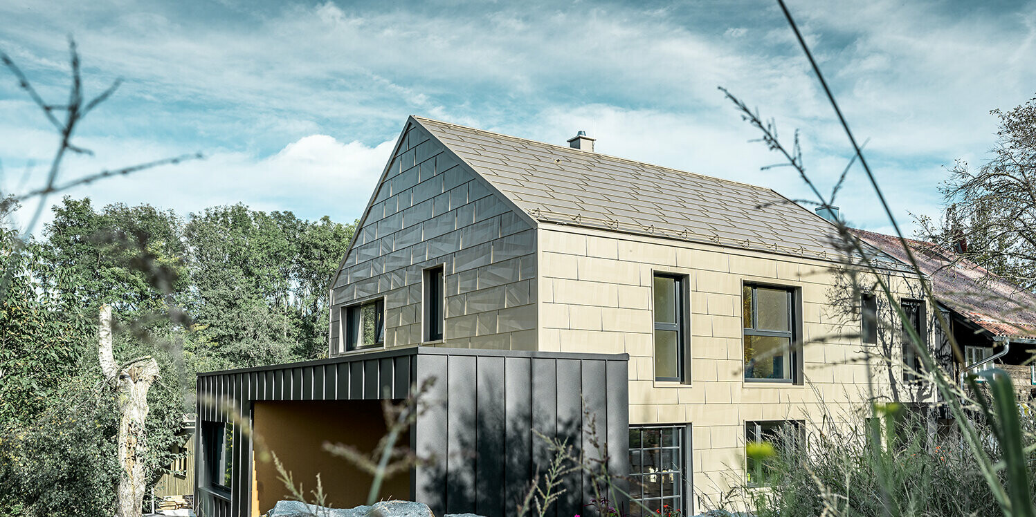 Apartment complex without eaves is clad on the roof and façade with the PREFA FX.12 aluminium panel in bronze. Right next to it is a garage with a flat roof with an angled standing seam façade.