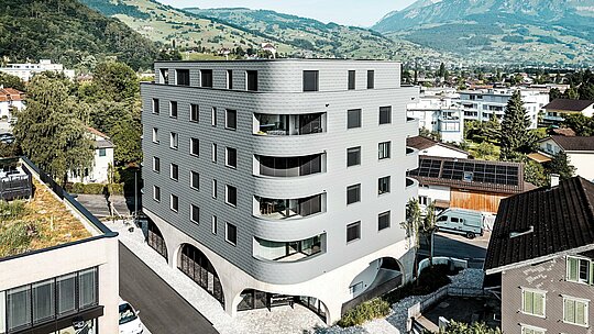 Luftaufnahme des Bernerhauses in Buchs mit markanter, dreieckiger Gebäudeform und weich gerundeten Balkonen inmitten der alpinen Landschaft. Die durchgängige Fassade aus PREFA Wandschindeln in P.10 Hellgrau umhüllt das Gebäude wie eine schuppenartige Aluminiumhaut und erzeugt ein lebendiges Spiel aus Licht und Struktur. Über dem massiven Betonsockel mit Arkaden wirkt die leichte, witterungsbeständige PREFA Aluminiumfassade besonders präzise, langlebig und wartungsarm – ein moderner Eyecatcher im Stadtbild.