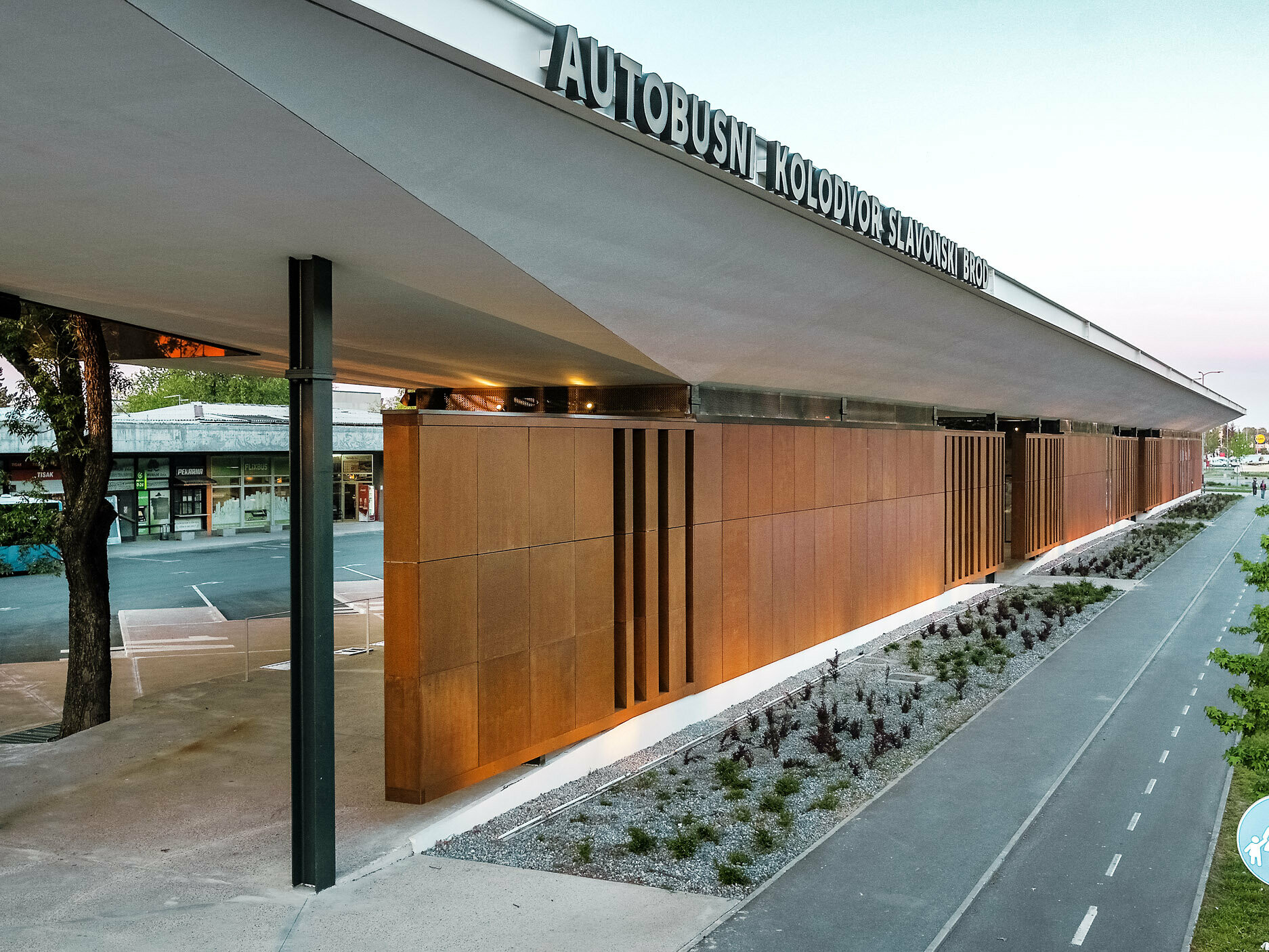 The picture shows the modern bus stop "Autobusni Kolodvor Slavonski Brod" in Croatia. The bus stop has a striking façade of brown panels supported by black columns. The white Prefalz roof from PREFA extends over the entire structure and provides protection for the areas below. A cycle path can be seen in the foreground, flanked by a well-tended green area and young trees. A light blue bus and various shops can be seen in the background. 