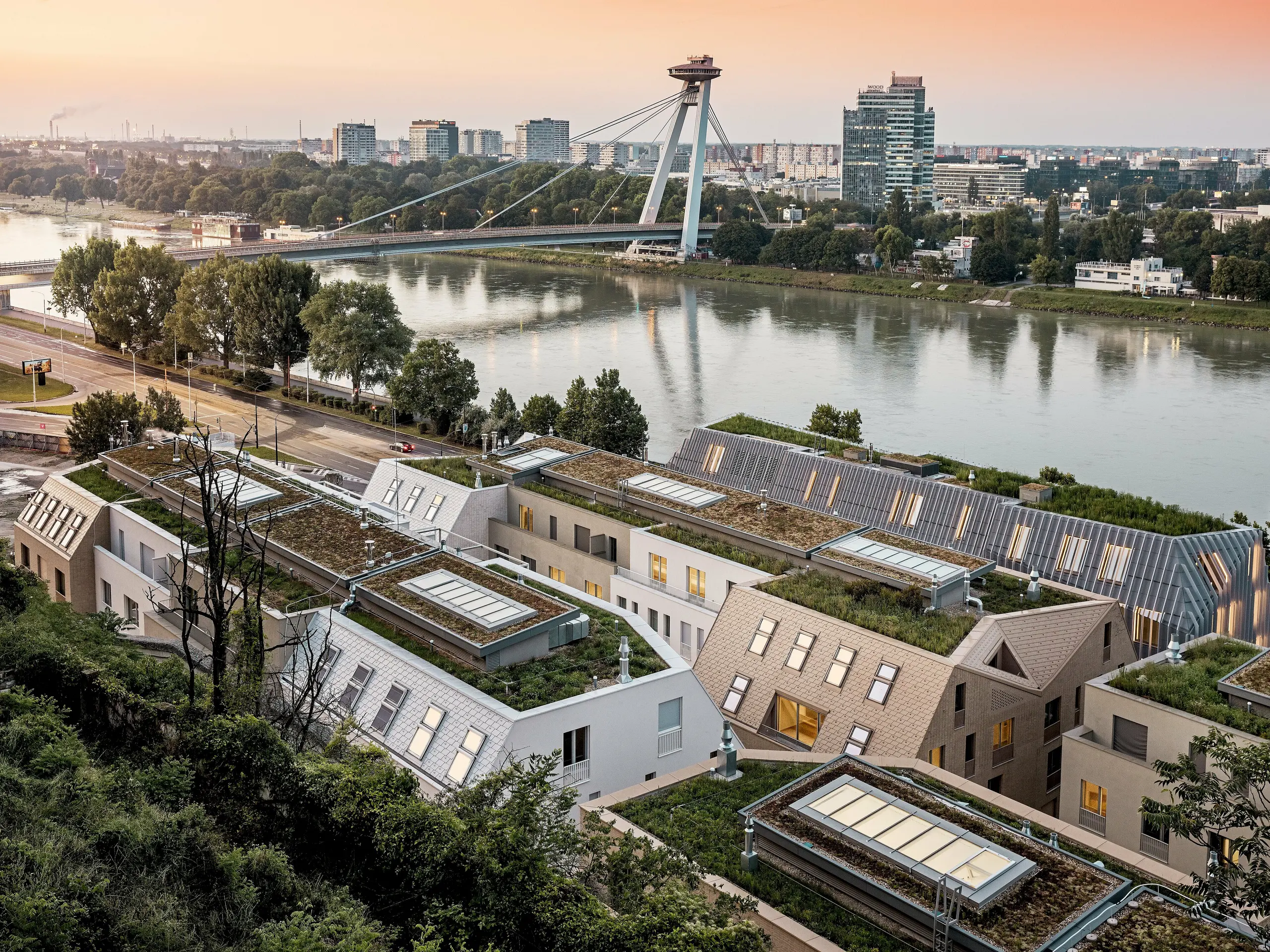 Spektakulärer Blick über das Stadtquartier Vydrica in Bratislava mit Donauufer und markanter Brücke im Abendlicht: Die vielschichtige Dachlandschaft mit begrünten Flächen, präzise integrierten Dachfenstern und steilen Satteldächern aus PREFA Aluminium Dachschindeln in Prefaweiß, Sandbraun und Bronze wird zum visuellen Highlight. Die klaren Linien und harmonischen Farbtöne verbinden Architektur und Umgebung eindrucksvoll. PREFA Aluminium überzeugt durch extreme Langlebigkeit, geringes Gewicht, hohe Witterungsbeständigkeit und flexible Gestaltung – ideal für komplexe, nachhaltige Dachlösungen.