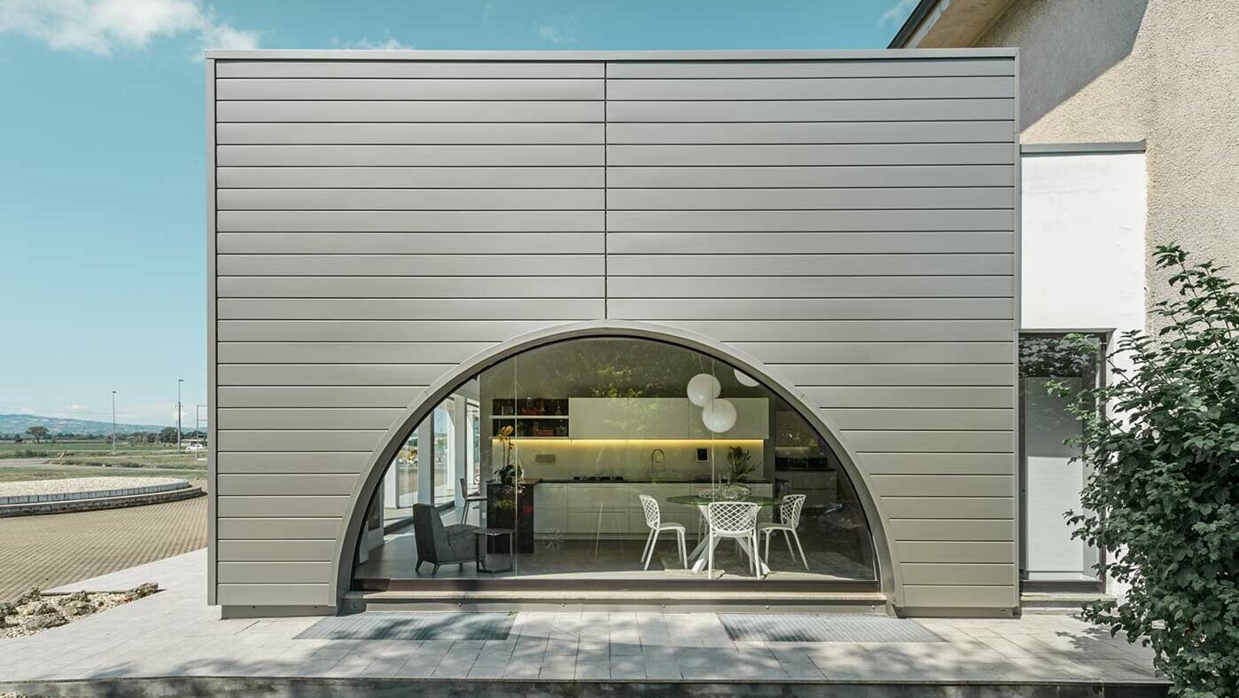 Flat-roof extension with a curved window façade, the façade is clad with horizontal aluminium panels, the PREFA sidings in smoke silver.