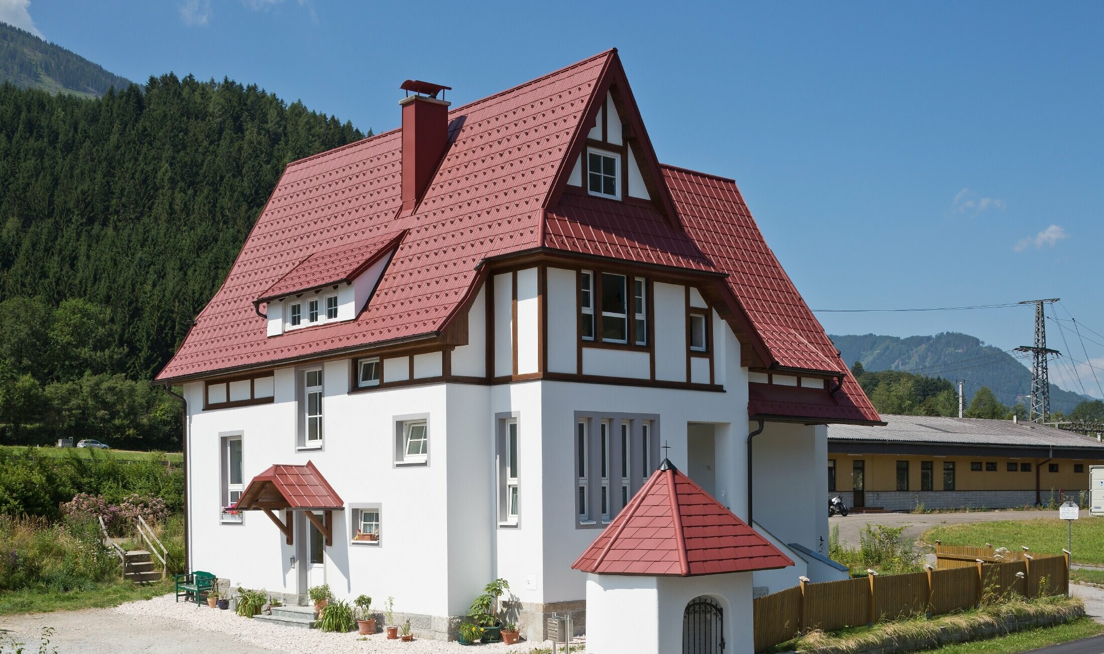 Steep, sloping roof with PREFA roof tiles in P.10 oxide red with many details such as dormers, turrets and chimney surrounds. The canopy and the wayside shrine in front of the house are also covered with PREFA sheet metal roofing.