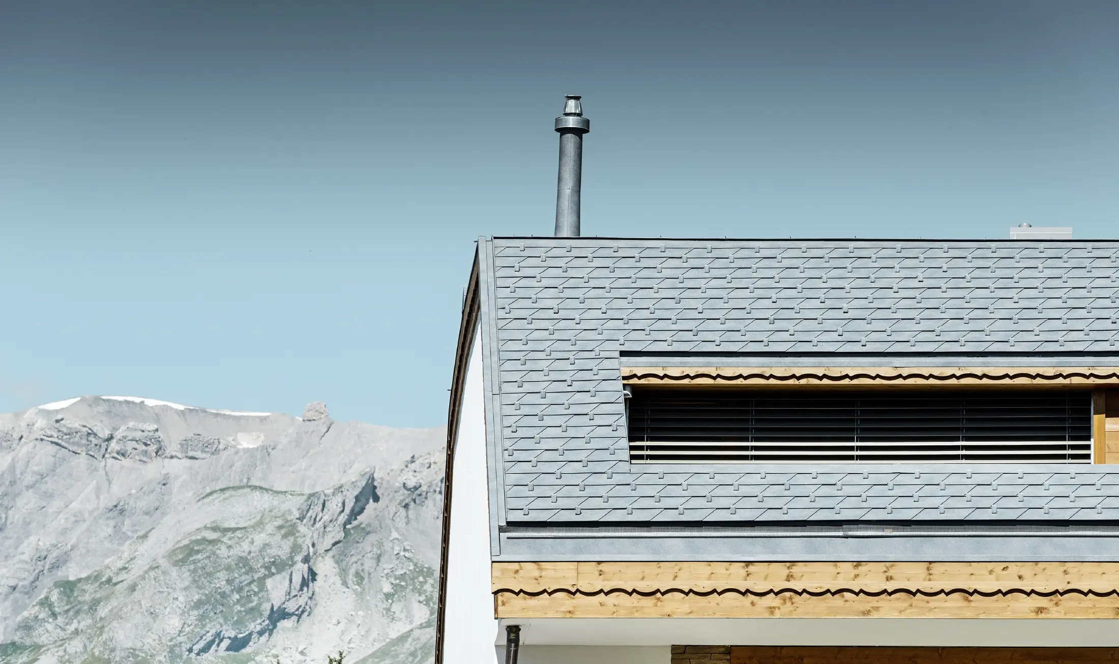 Apartment building in Crans-Montana (Switzerland) with mountains in the background, a façade with lovely wooden elements and a PREFA aluminium shingle roof in stone grey