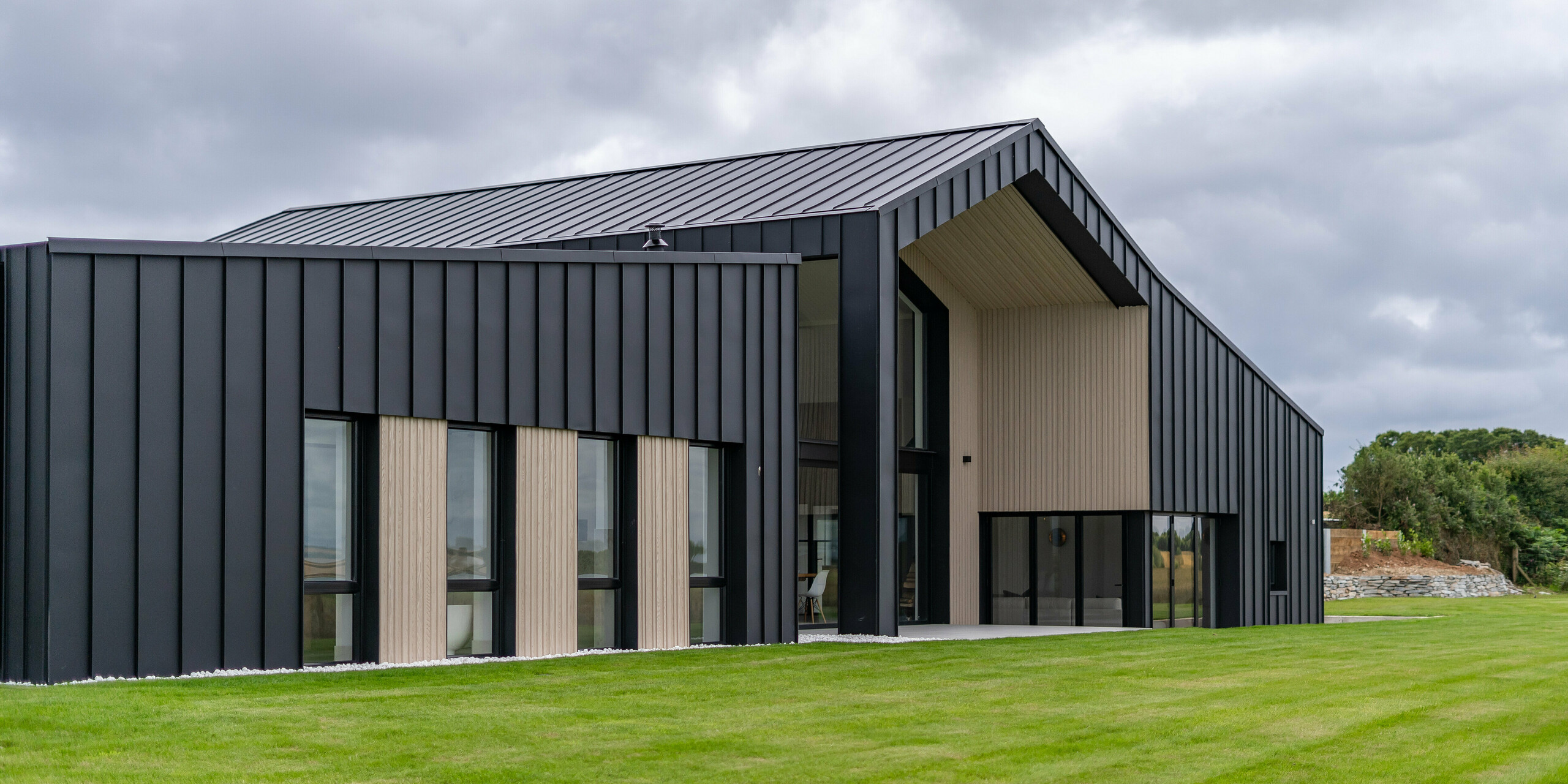 An oblique view of the rear of the detached house ‘The Hide’ in Cornwall, emphasised by the PREFALZ aluminium facade in P.10 anthracite. The precise standing seam covering made of high-quality metal sheets forms an elegant contrast to the wooden elements, while the generous roof with its clear lines underlines the modern architecture of the building. Surrounded by well-kept greenery, the design blends harmoniously into the landscape.