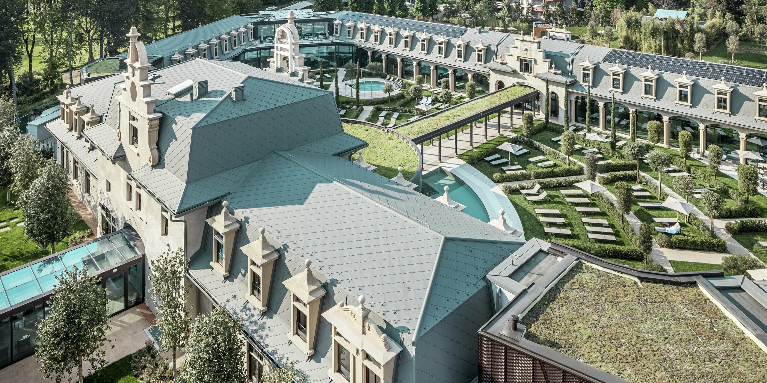 Panoramablick auf den vollständig sanierten Thermenkomplex „De Montel – Terme Milano“ mit großzügiger Gartenanlage, geschwungenen Wegen und Liegebereichen rund um zahlreiche Thermalbecken. Die Dächer sind durchgängig mit der PREFA Dachraute 44 × 44 in P.10 Hellgrau eingedeckt, ergänzt durch nahtlos integrierte Photovoltaikmodule und begrünte Flächen. Historische Architektur und nachhaltige Materialien verbinden sich zu einem innovativen, urbanen Erholungsraum.