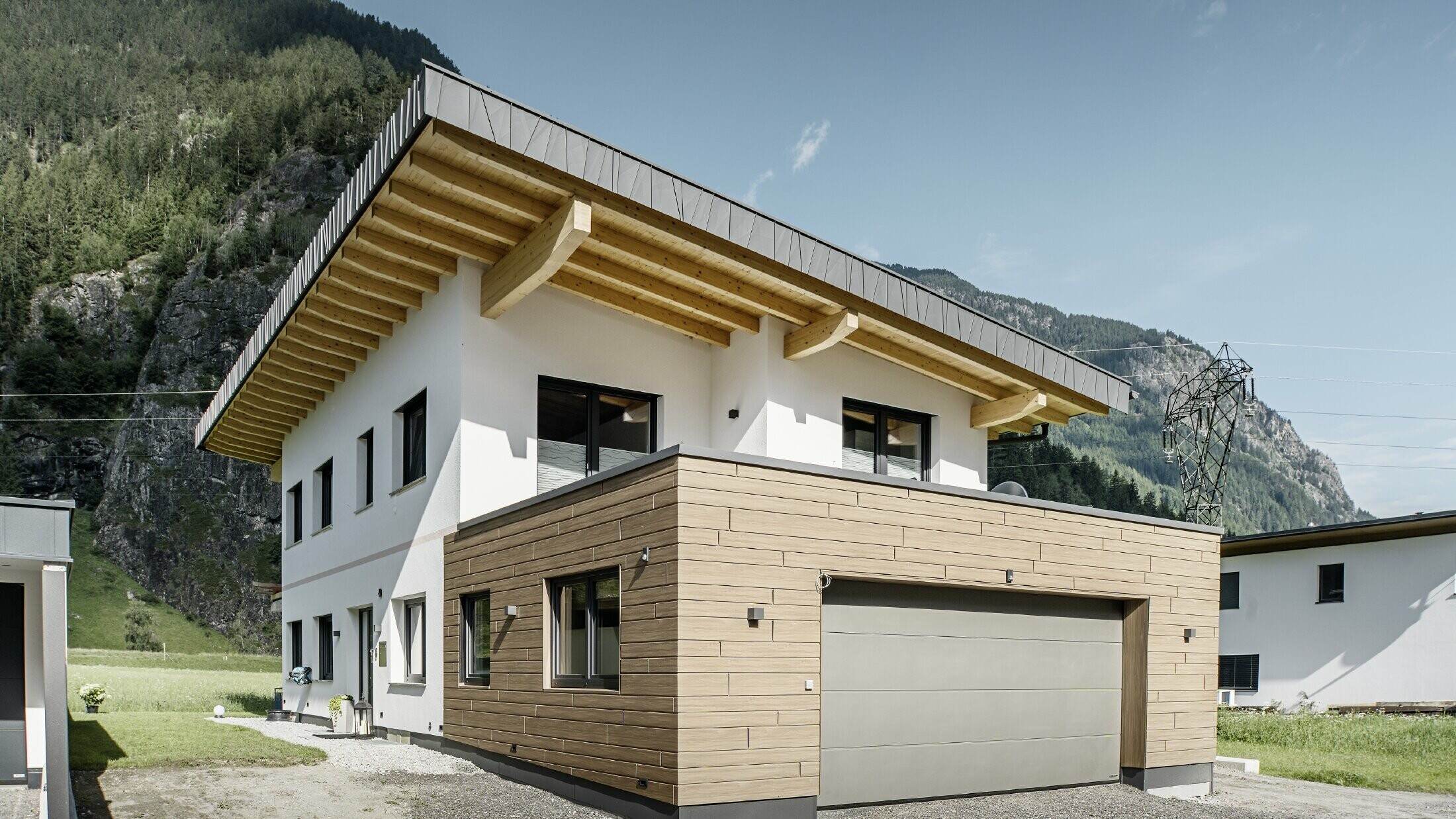 A modern detached house with a refurbished façade: a renewed exterior façade featuring horizontal cladding with a wood-effect finish, combined with white-plastered surfaces, integrated garage cladding and contemporary architecture set in an alpine setting.