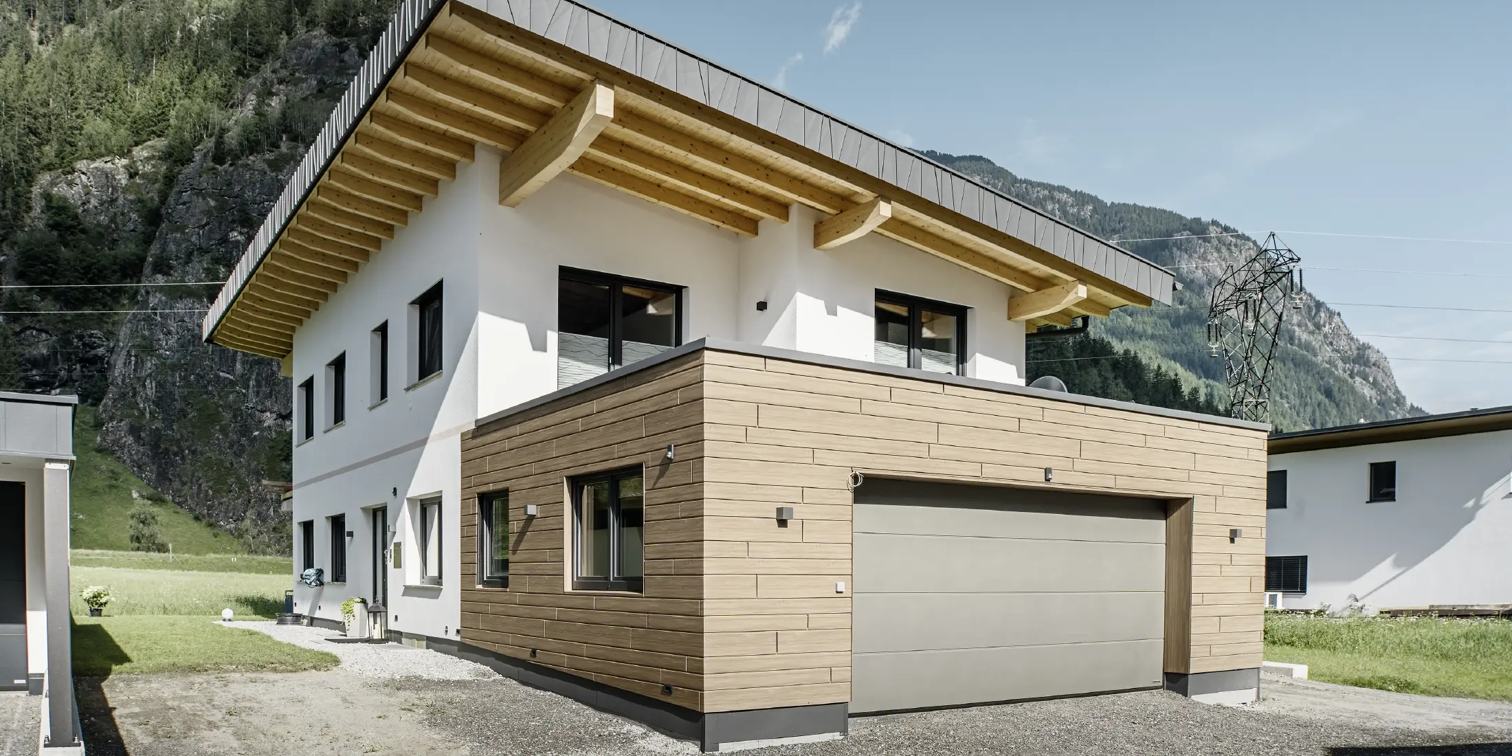 A modern detached house with a refurbished façade: a renewed exterior façade featuring horizontal cladding with a wood-effect finish, combined with white-plastered surfaces, integrated garage cladding and contemporary architecture set in an alpine setting.