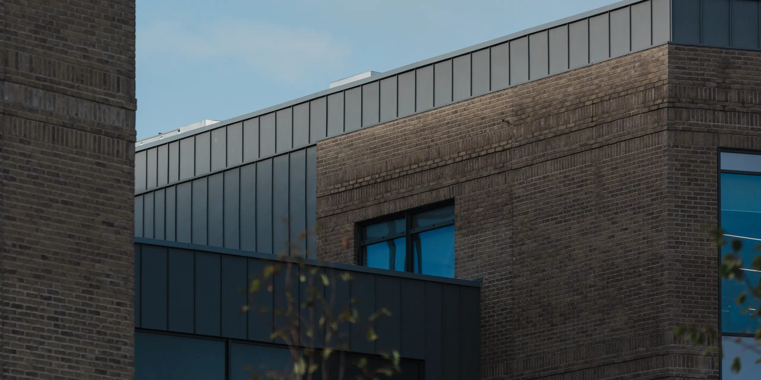 A close-up architectural view of the Northern Regional College in Ballymena highlights the upper façade edge finished with vertical PREFALZ standing seam aluminium panels in patina grey. The crisp aluminium band creates a strong contrast to the textured brickwork and recessed windows beneath. Set against a blue sky, the smooth metal surface subtly reflects daylight and reinforces the building’s clean geometry. PREFA aluminium delivers durability, precise detailing and a timeless, low-maintenance façade solution for contemporary educational architecture.