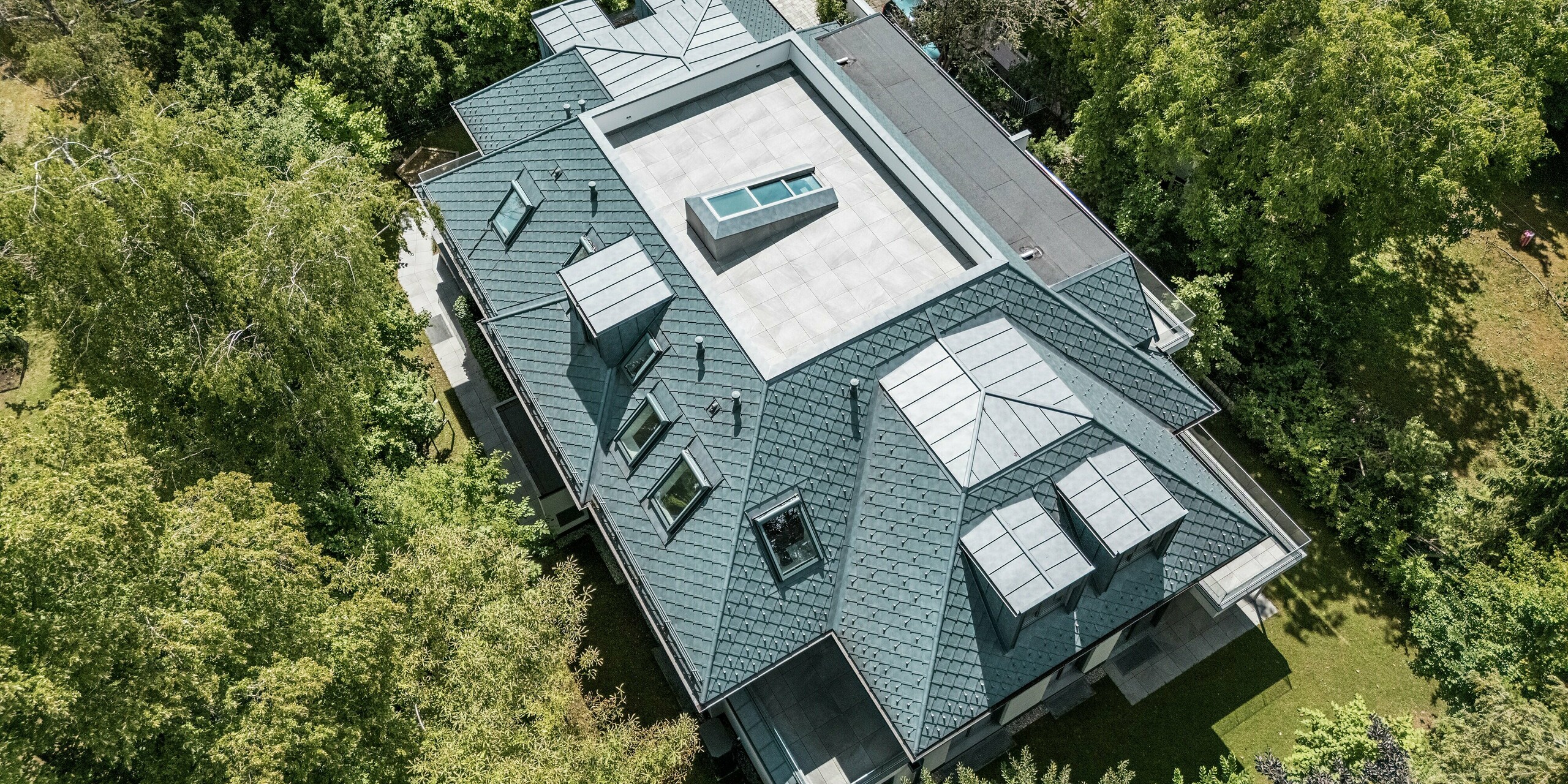 Diese Luftaufnahme zeigt ein modernes Mehrfamilienhaus mit einem markanten Mansardflachdach, das mit PREFA Dachrauten in der Größe 29 × 29 und der Farbe P.10 Steingrau gedeckt ist. Das Dachdesign wird durch Flachdachgauben aus PREFALZ ergänzt und sorgt so für eine markante, strukturierte Optik, die sich perfekt in die grüne Umgebung einfügt. Der zentrale, begehbare Flachdachbereich verstärkt die moderne Architektur. Die Verwendung langlebiger und witterungsbeständiger Aluminiumprodukte garantiert ästhetische und funktionale Vorteile.