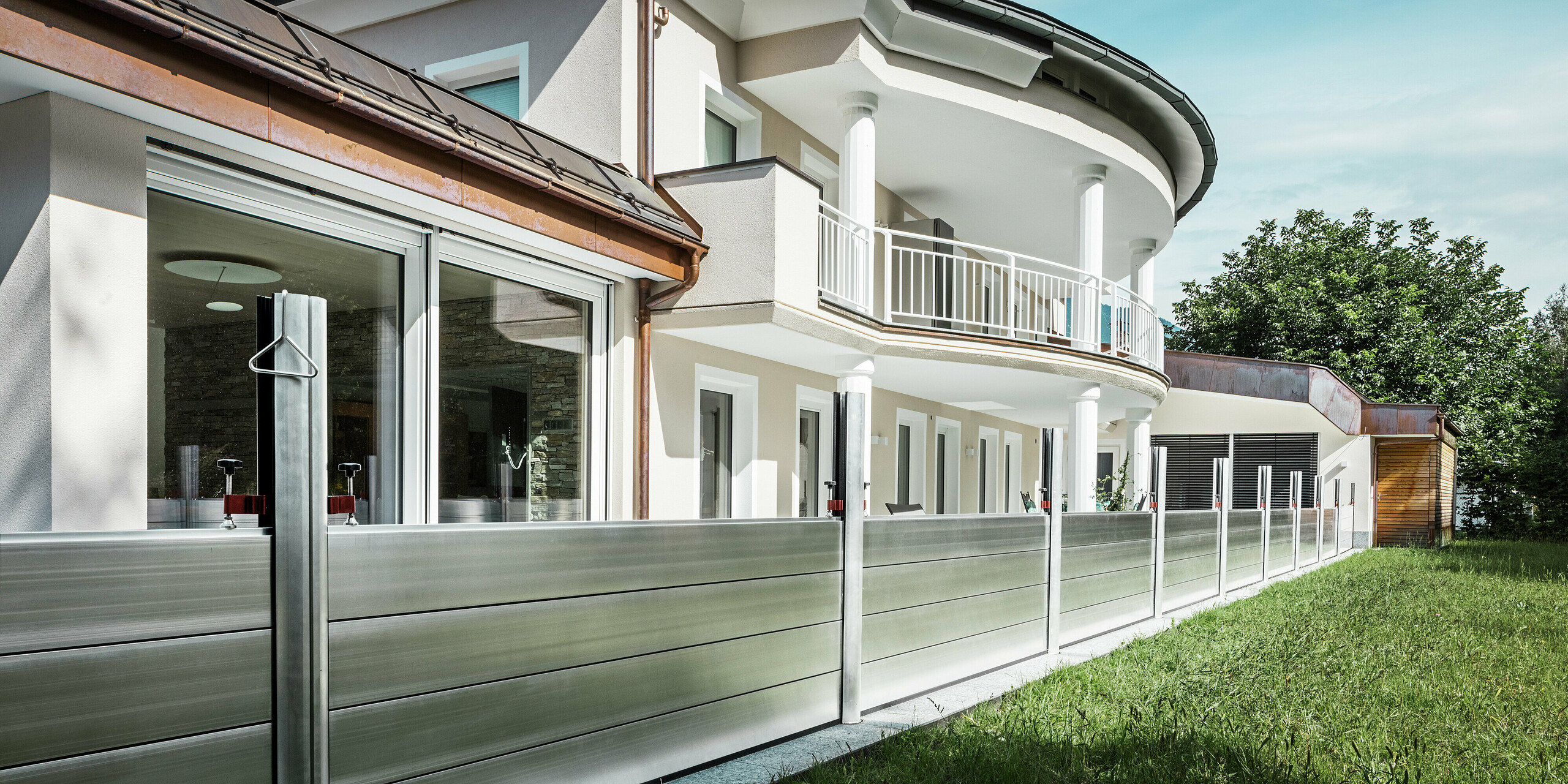 Blick auf ein modernes Wohnhaus in Tirol mit installiertem PREFA Hochwasserschutzsystem entlang der Terrasse. Die robusten Dammbalken aus Aluminium sind zwischen stabilen Stützprofilen montiert und schützen das Gebäude vor Überflutungen aus der angrenzenden Grünfläche mit Gewässern. Das elegante Haus mit Rundbalkon, weißen Säulen und großflächigen Fenstern zeigt, wie sich funktioneller Hochwasserschutz harmonisch in hochwertige Architektur integrieren lässt.