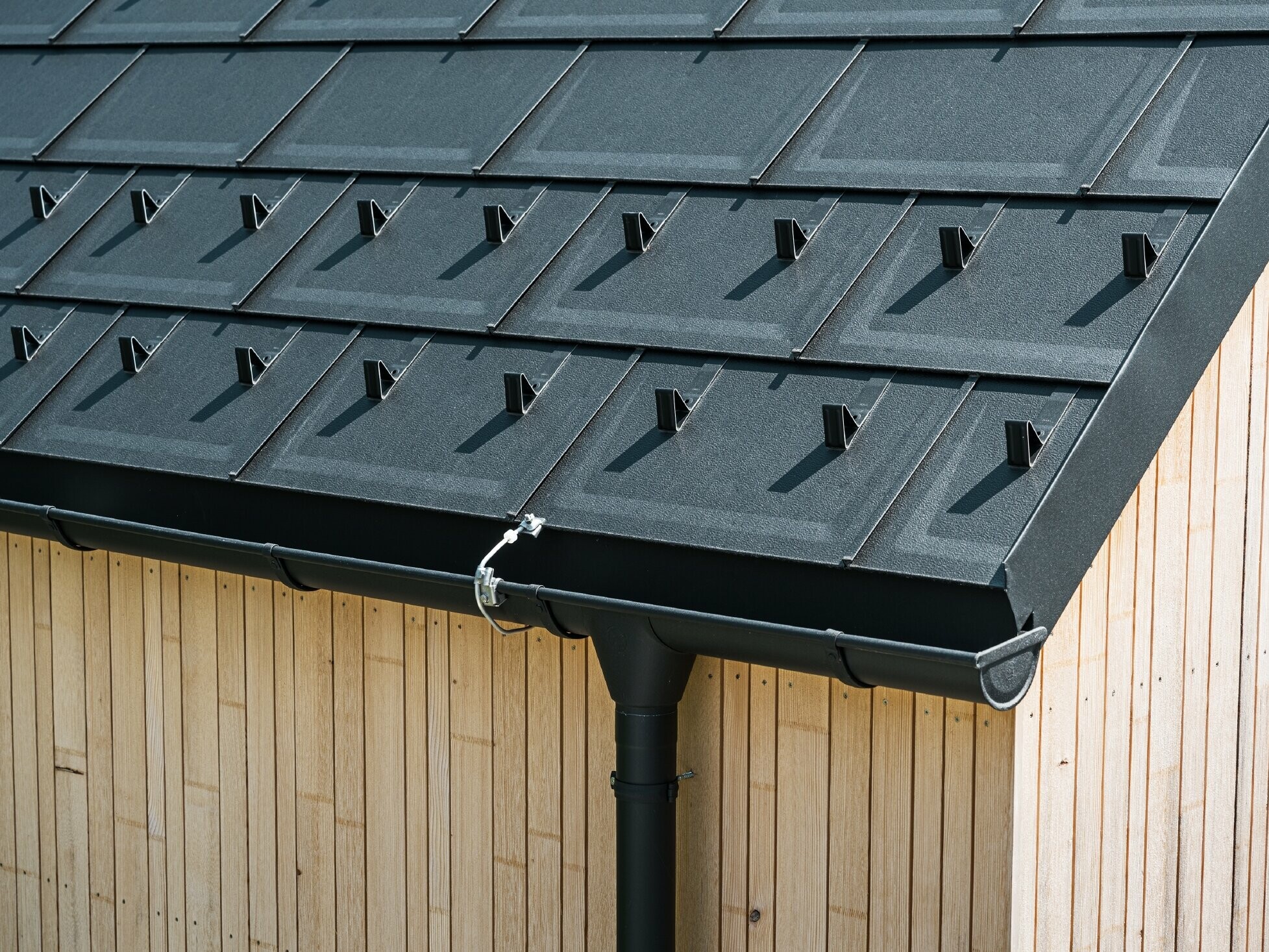 Close-up of PREFA roof tile R.16 with snow stoppers, gutter and downpipe in black P.10 incl. lightning protection and a vertical wooden façade