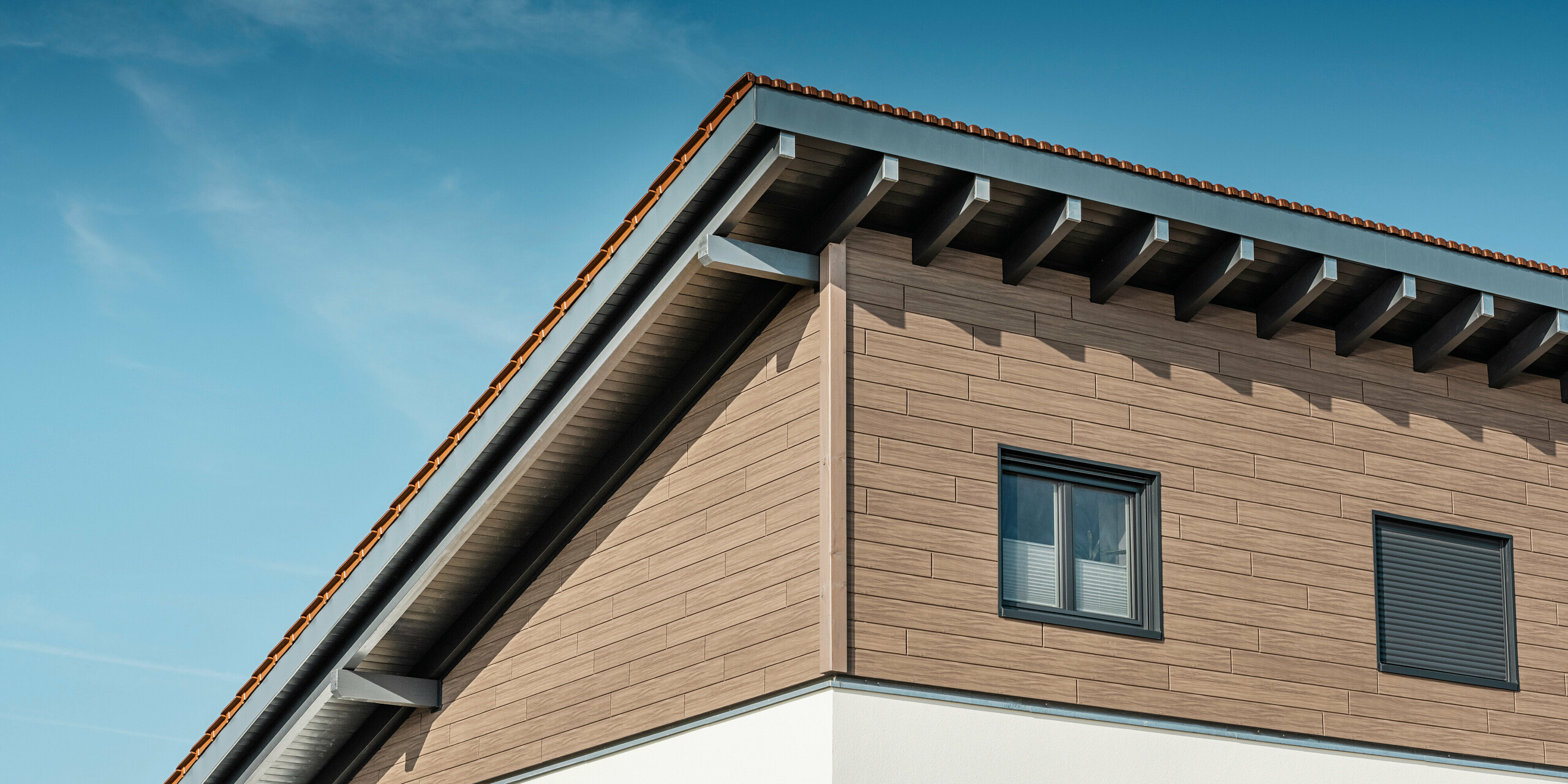 Schräger Blick von unten auf das Obergeschoss eines Einfamilienhauses in Bruck. Das obere Stockwerk ist mit PREFA Sidings in einer Holzoptik umhüllt. Zu sehen ist das Pultdach, das auf der Gebäuderückseite nach unten verläuft.