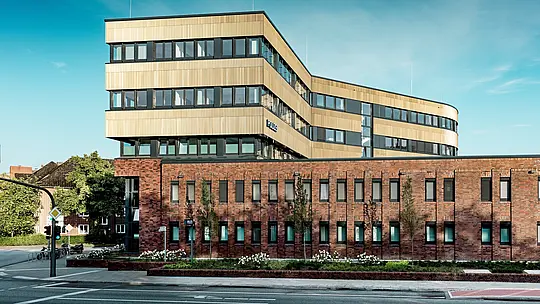 Weitläufige Ansicht des Polizeikommissariats 43 in Hamburg: Über dem robusten Backsteinbestand erhebt sich der moderne Erweiterungsbau mit einer markanten PREFA Aluminiumfassade aus Strangpressprofilen „Zacke“ in Mittelbronze. Die fein strukturierte Oberfläche erzeugt lebendige Lichtreflexe und betont die klare, horizontale Gliederung sowie die sanfte Rundung des Baukörpers. Als prägnanter Eyecatcher vereint die Fassade architektonische Eleganz mit den Vorteilen von PREFA Aluminium – langlebig, korrosionsbeständig und nahezu wartungsfrei.