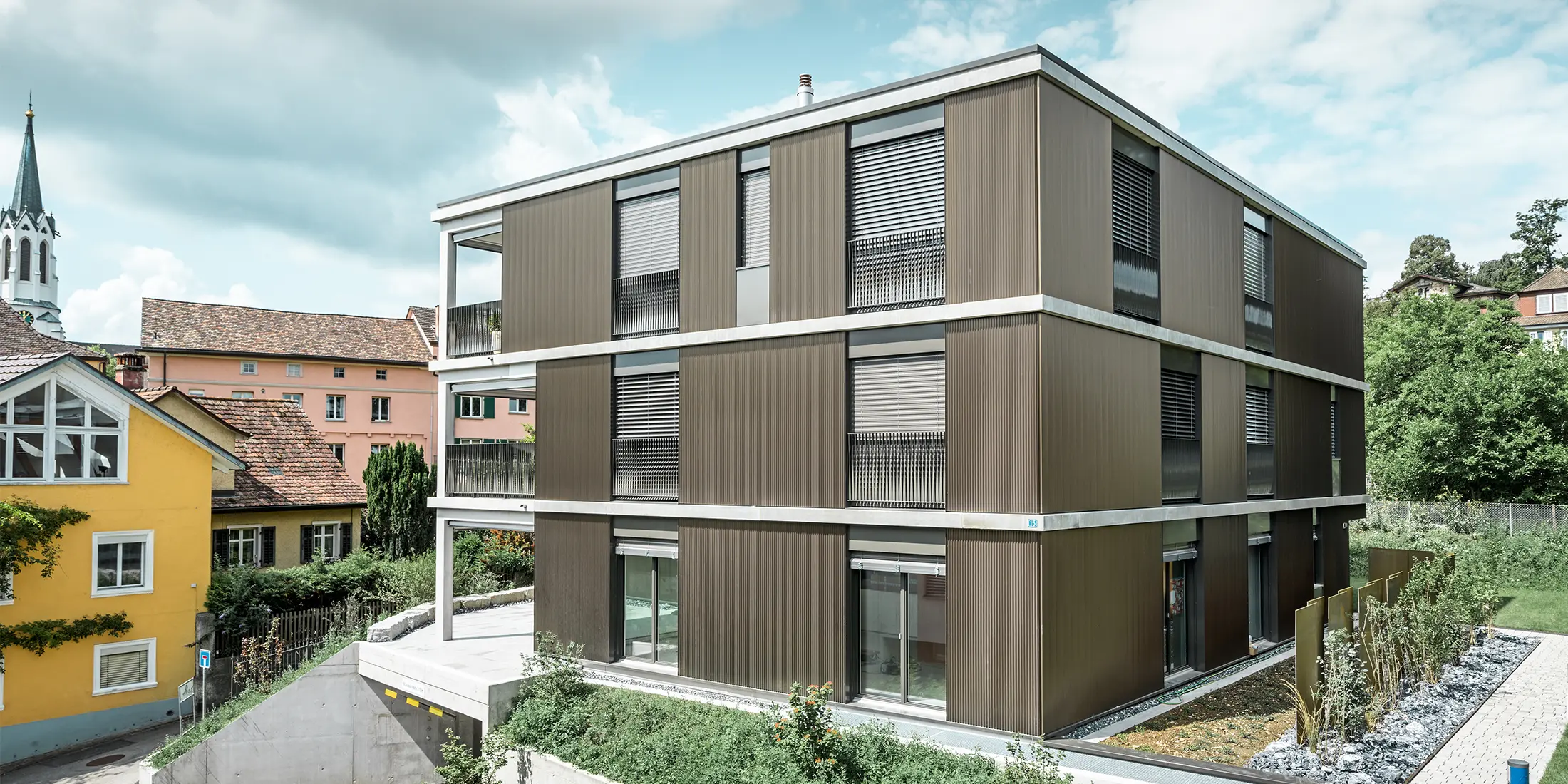 The striking aluminium façade of this apartment building in Schaffhausen features the elegant PREFA serrated profile in the colour Colinal. The vertical arrangement of the extruded aluminium profiles makes the building appear more compact and gives the façade structure a harmonious balance. The sophisticated design blends tastefully into the surroundings and gives the building a modern elegance that is both visually appealing and functional. The perspective shows the apartment building from the front with the garage entrance. Trees, other buildings and a church tower can be seen in the background.