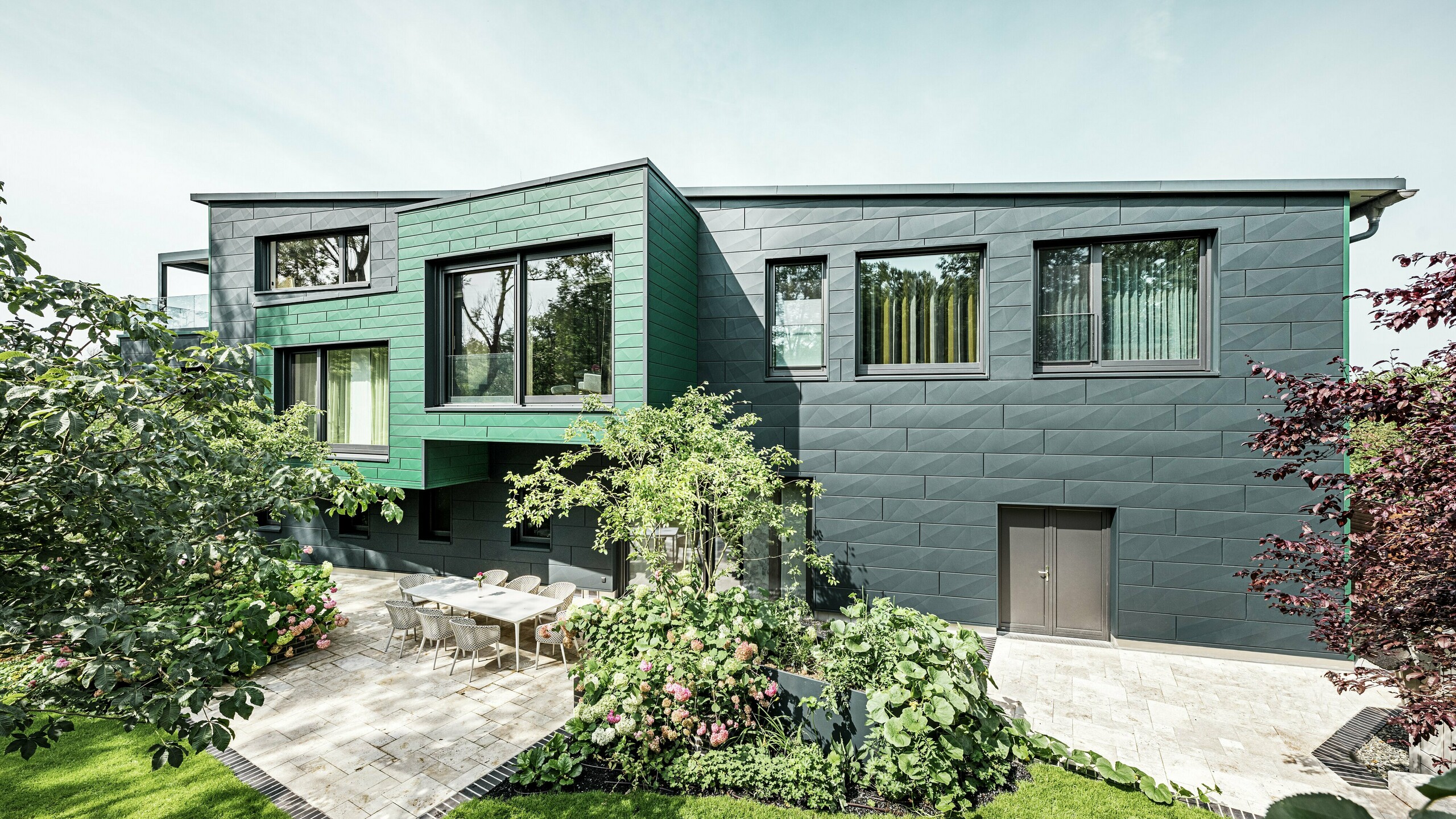 The detached house in Türkheim impresses with its unusual façade, which is clad with PREFA Siding X panels in anthracite and moss green. The clean lines and striking colour choice of the aluminium panels give the building a modern and elegant look. In the foreground is the spacious garden area with a stylish terrace and a dining table with modern chairs. Colourful flower beds and healthy plants provide a lively, green contrast to the robust façade. The durable and low-maintenance aluminium façade protects the building and ensures timeless beauty.