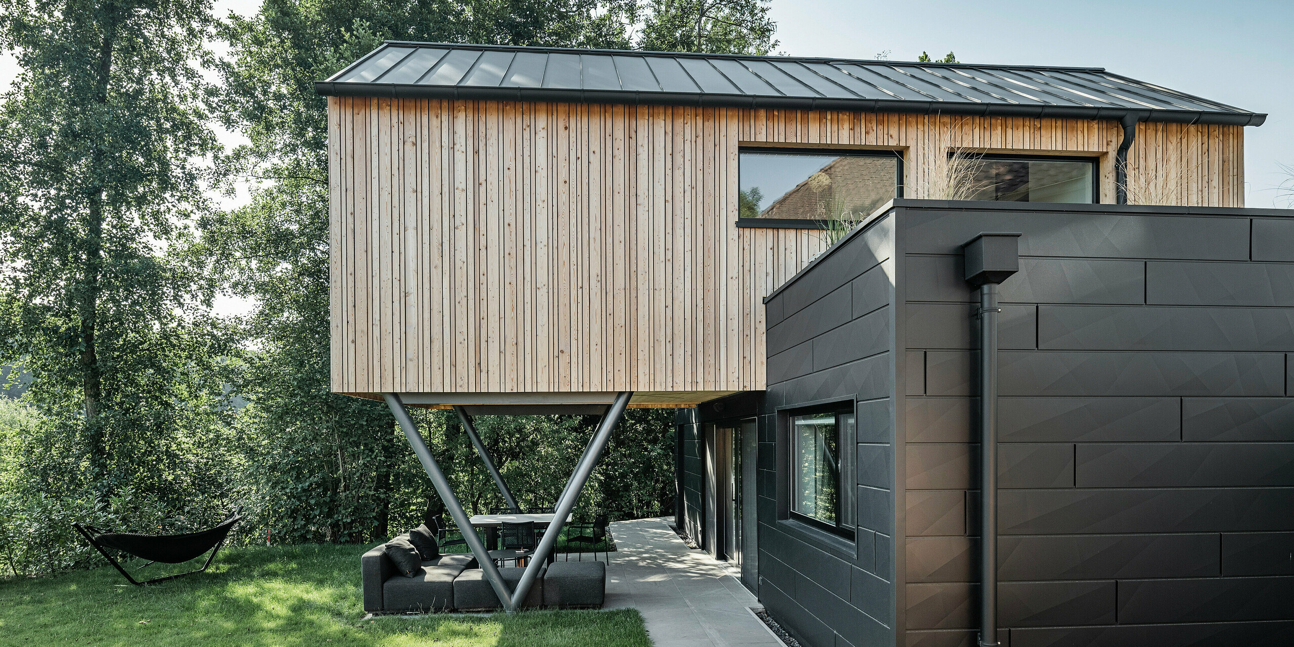 Das 'Woodstock Chalet 5' präsentiert sich mit einer Symbiose aus moderner Architektur und natürlicher Umgebung, akzentuiert durch die PREFA Siding.X Fassadenelemente in P.10 Anthrazit. Diese Aluminiumelemente verleihen der unteren Gebäudeebene eine markante Textur und Farbtiefe, die sich vom hölzernen Obergeschoss abhebt. Die großzügigen Fensterflächen bieten einen Blick auf die umliegende Grünlandschaft, während die metallenen Stützträger der überhängenden Konstruktion ein industriell-modernes Flair einführen.