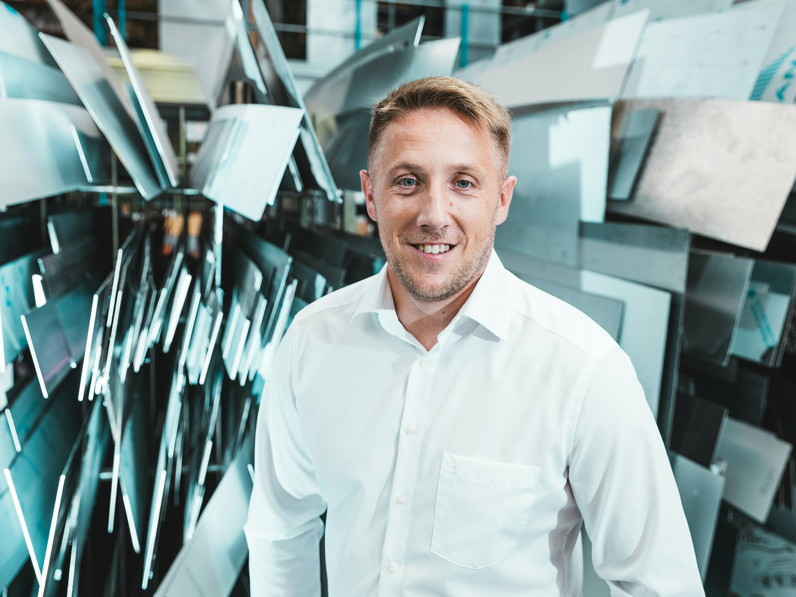 Selbstbewusst und freundlich steht Projektleiter Marcel Schmidt von der Spenglerei ASA Schüßler vor einer eindrucksvollen Wand aus unterschiedlich geformten Aluminiumelementen. Die dynamisch arrangierten Metallplatten spiegeln das Licht effektvoll wider und symbolisieren Innovation, Präzision und Qualität in der Metallverarbeitung. Der Mann im weißen Hemd verkörpert Kompetenz und moderne Fertigungsexzellenz. Im Hintergrund wird die vielfältige Verarbeitungsmöglichkeit von Aluminium sichtbar – von Dach- und Fassadensystemen bis hin zu maßgefertigten Sonderlösungen. 