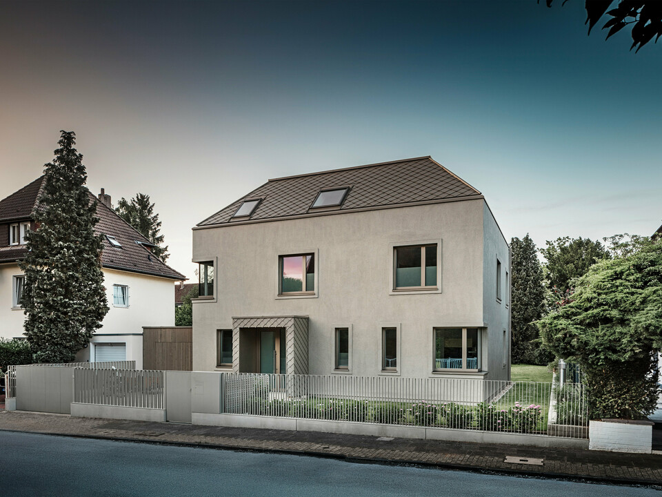 A modern detached house with a hipped roof and bronze-coloured aluminium roof and façade cladding from PREFA. The house is located in a quiet residential neighbourhood, surrounded by well-tended gardens and other detached houses. A low fence separating the property from the street can be seen in the foreground. The architecture of the house is characterised by clean lines and large windows that allow plenty of natural light into the interior.