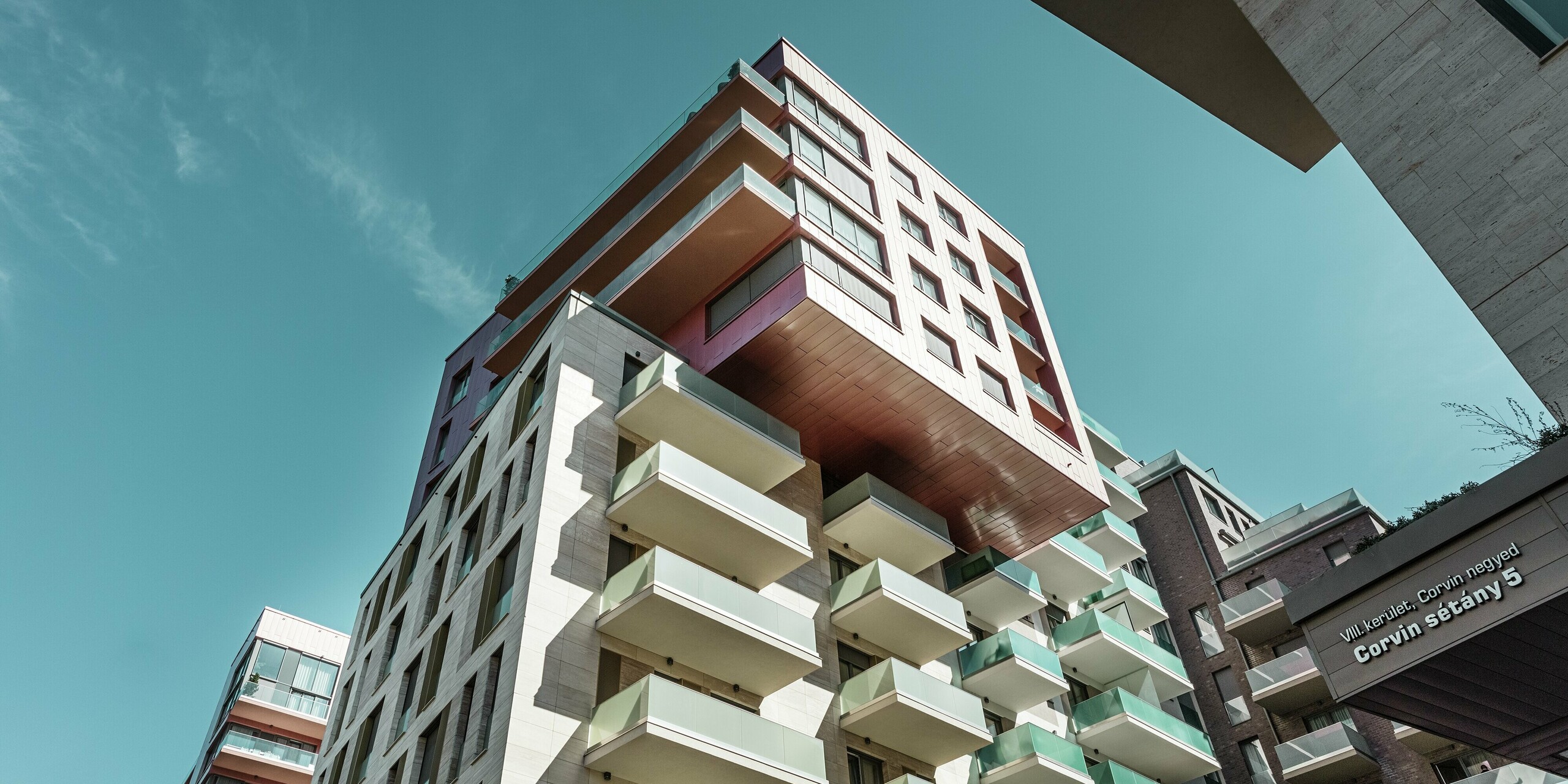Markante Froschperspektive auf den Grand Corvin Wohnkomplex im Corvin-Viertel von Budapest mit auskragendem Gebäudeteil, der vollständig mit PREFA Siding Fassadenpaneelen in Sonderfarbe Lachsmetallic verkleidet ist. Die vertikale Verlegung der langlebigen Aluminiumfassade verleiht dem modernen Stadtbaukörper eine elegante Struktur und hebt die hochwertige Architektur im Rahmen der groß angelegten Stadterneuerung wirkungsvoll hervor.