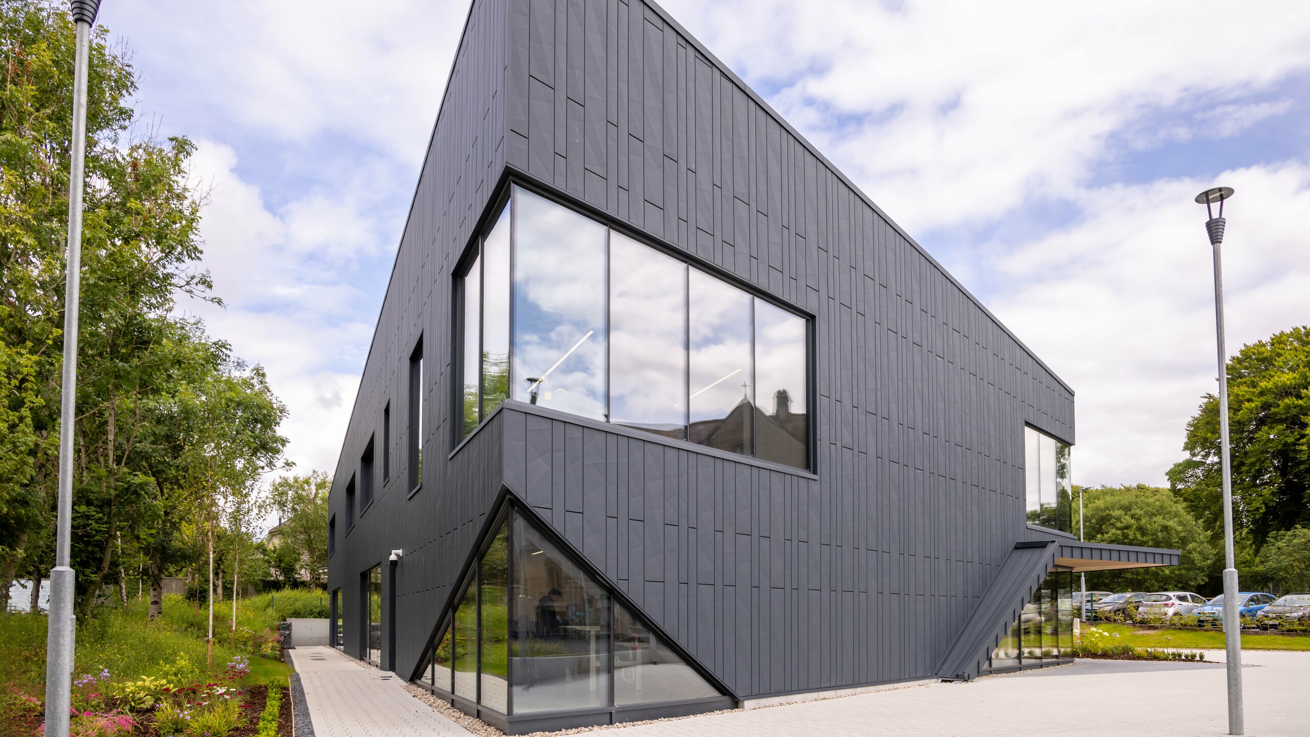 View of the front of the CREW building in Galway, Ireland, with a modern façade made of PREFA Siding.X façade panels and PREFALZ in the colour P.10 anthracite. The vertical lines of the façade and the large window areas emphasise the innovative and dynamic design of the building. The aluminium cladding blends harmoniously into the surrounding landscape and underlines the high-quality architecture of the building.