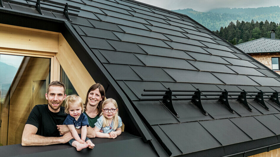 The two homeowners and their children are enjoying the view from the loggia of their newly built house, which is fitted with stylish PREFA solar roof panels in P.10 black.