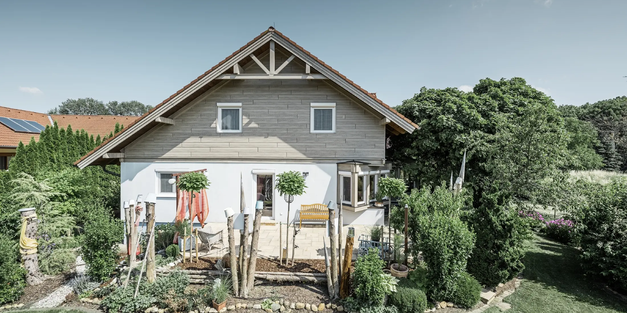A detached house with a refurbished façade, featuring new gable cladding in a modern wood-effect finish in grey-beige, combined with a rendered façade, a gabled roof and a well-tended garden.
