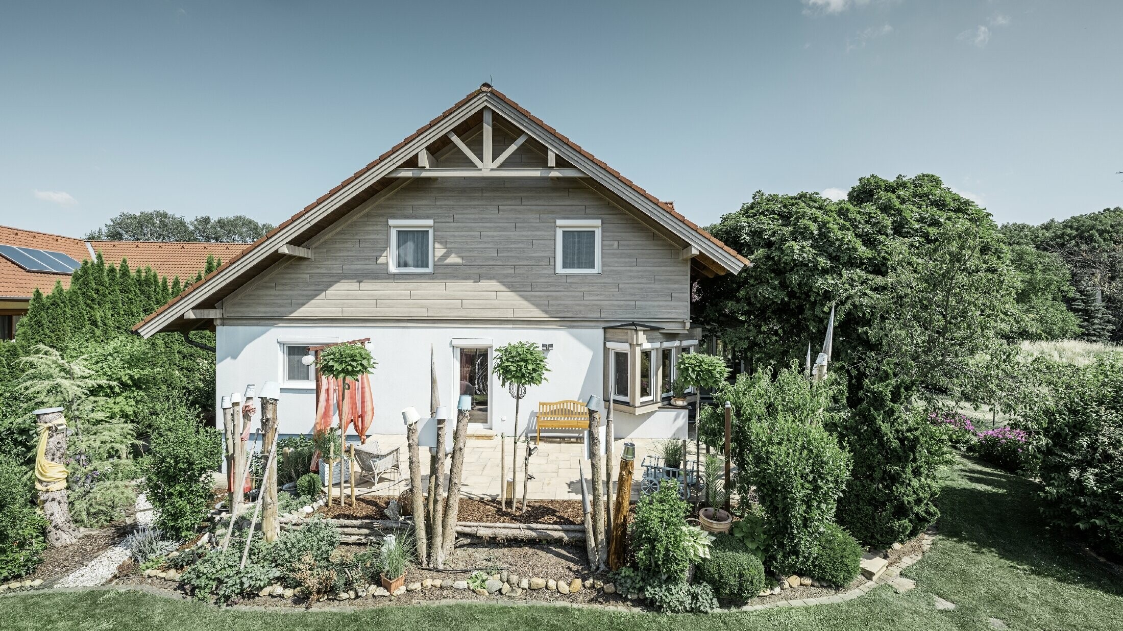 A detached house with a refurbished façade, featuring new gable cladding in a modern wood-effect finish in grey-beige, combined with a rendered façade, a gabled roof and a well-tended garden.
