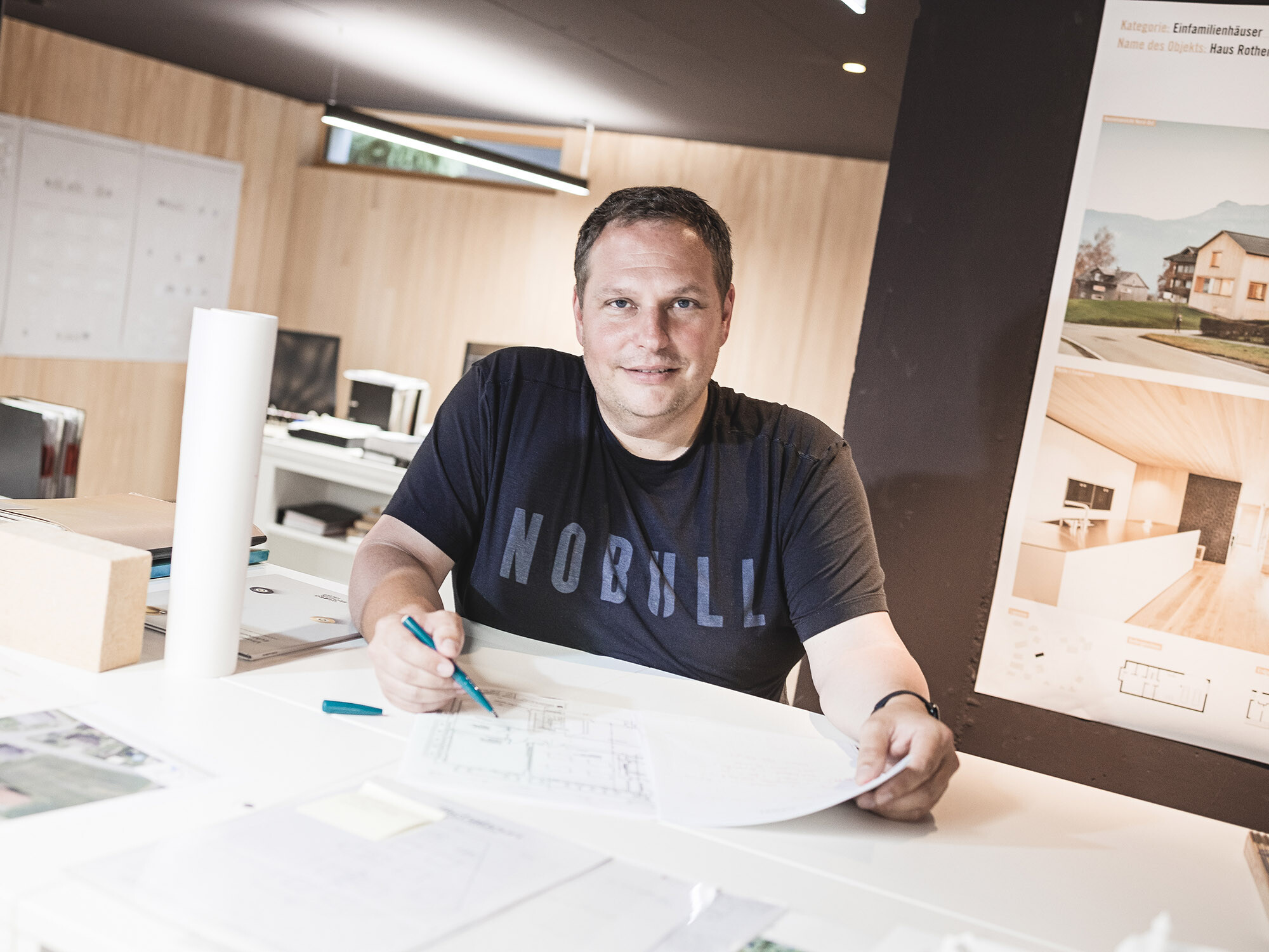 Portrait of the lead builder Jürgen Haller at his office desk, holding a pen in his one hand, a plan in his other hand.