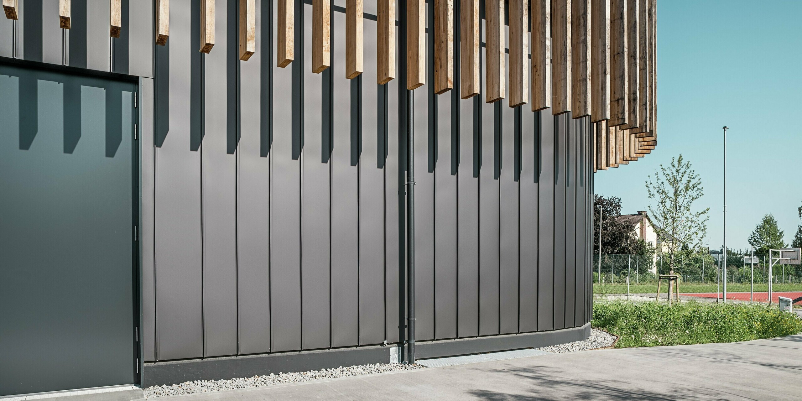 Nahaufnahme des Erdgeschosses der Team 7 Welt in Ried im Innkreis: Die schwarzbraune PREFALZ Fassade bildet mit den vertikalen Holzelementen ein harmonisches Gesamtbild. Die robuste Aluminiumverkleidung schützt vor Witterungseinflüssen und überzeugt durch ihre langlebige, wartungsfreie Qualität. Das Spiel von Licht und Schatten unterstreicht die klaren Linien und die moderne Architektur des Gebäudekomplexes.