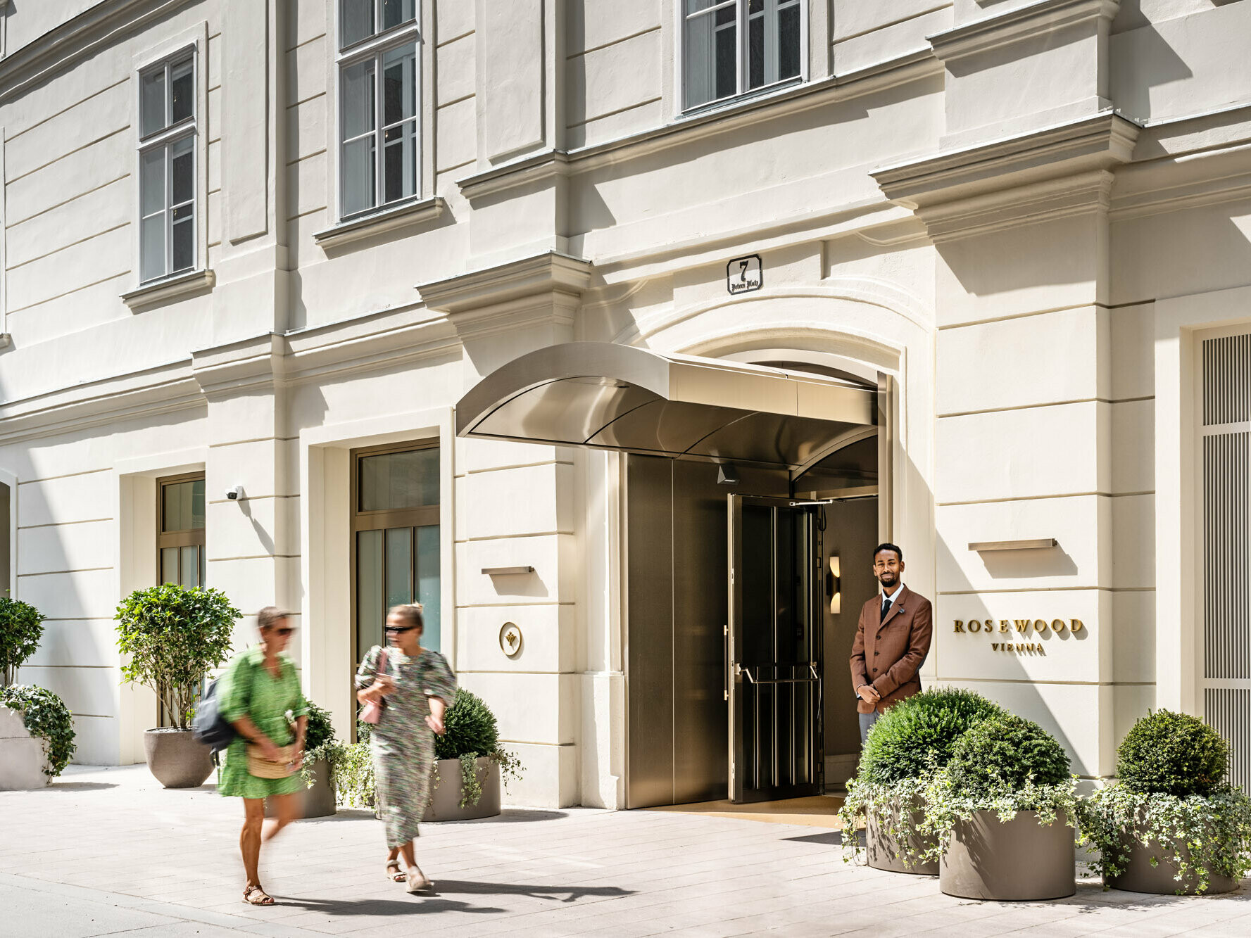 Entrance to the Rosewood Hotel with a porter, passers-by and larger potted plants in front of it.