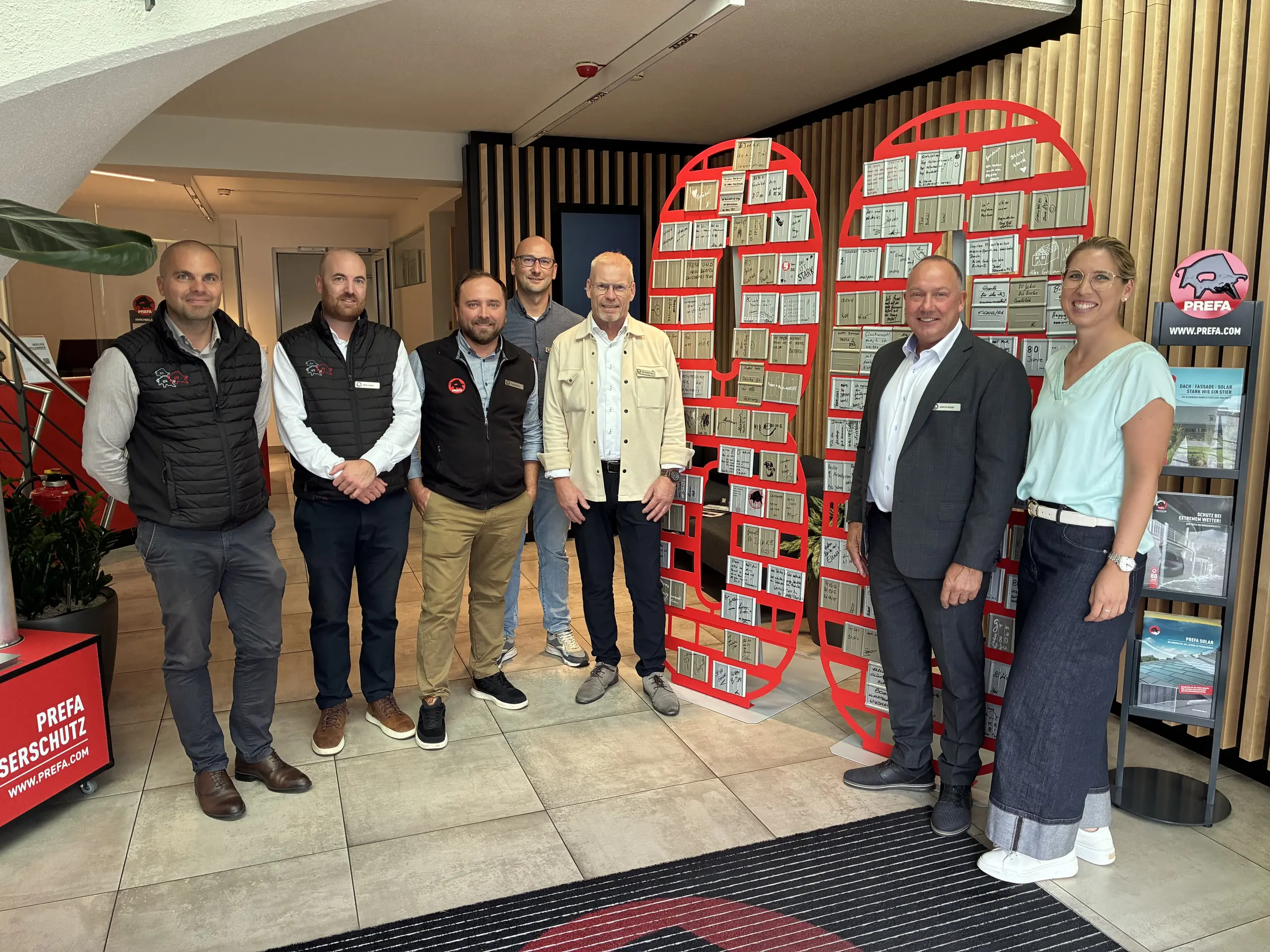 A group photo of six people indoors in front of two red display panels featuring numerous metal samples.