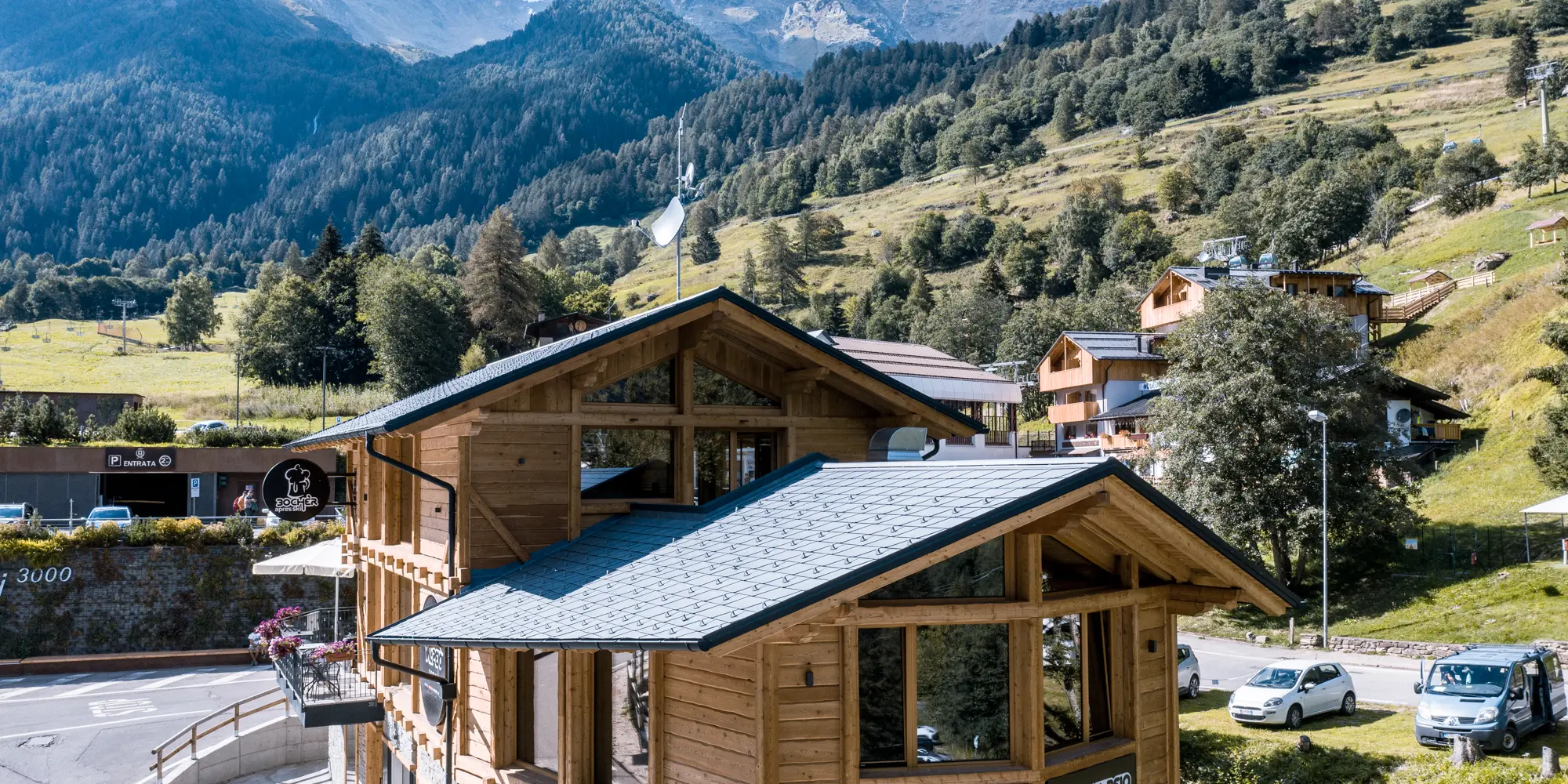 Nahaufnahme eines charmanten, hölzernen Après-Ski-Lokals in einer alpinen Landschaft, gekrönt von PREFA Aluminiumdachschindeln DS.19 in P.10 Steingrau. Der malerische Bau bietet eine Terrasse für Gäste, die eine atemberaubende Aussicht auf die italienischen Berge genießen können, wobei das Lokal nahtlos in die umliegende Architektur und die üppigen Bergwiesen einfließt.