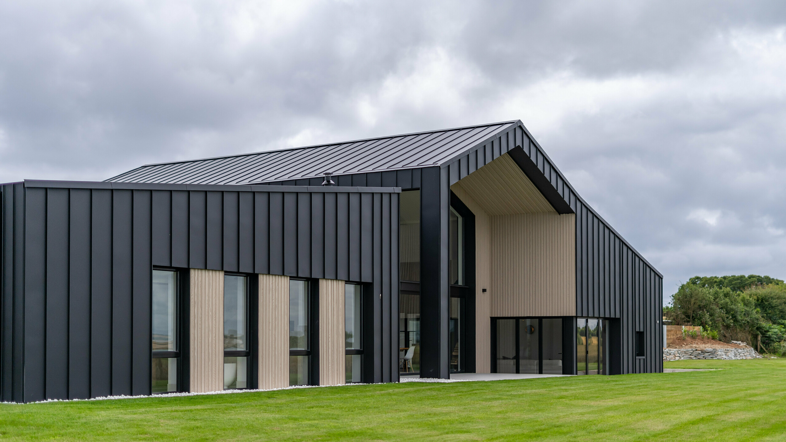 An oblique view of the rear of the detached house ‘The Hide’ in Cornwall, emphasised by the PREFALZ aluminium facade in P.10 anthracite. The precise standing seam covering made of high-quality metal sheets forms an elegant contrast to the wooden elements, while the generous roof with its clear lines underlines the modern architecture of the building. Surrounded by well-kept greenery, the design blends harmoniously into the landscape.