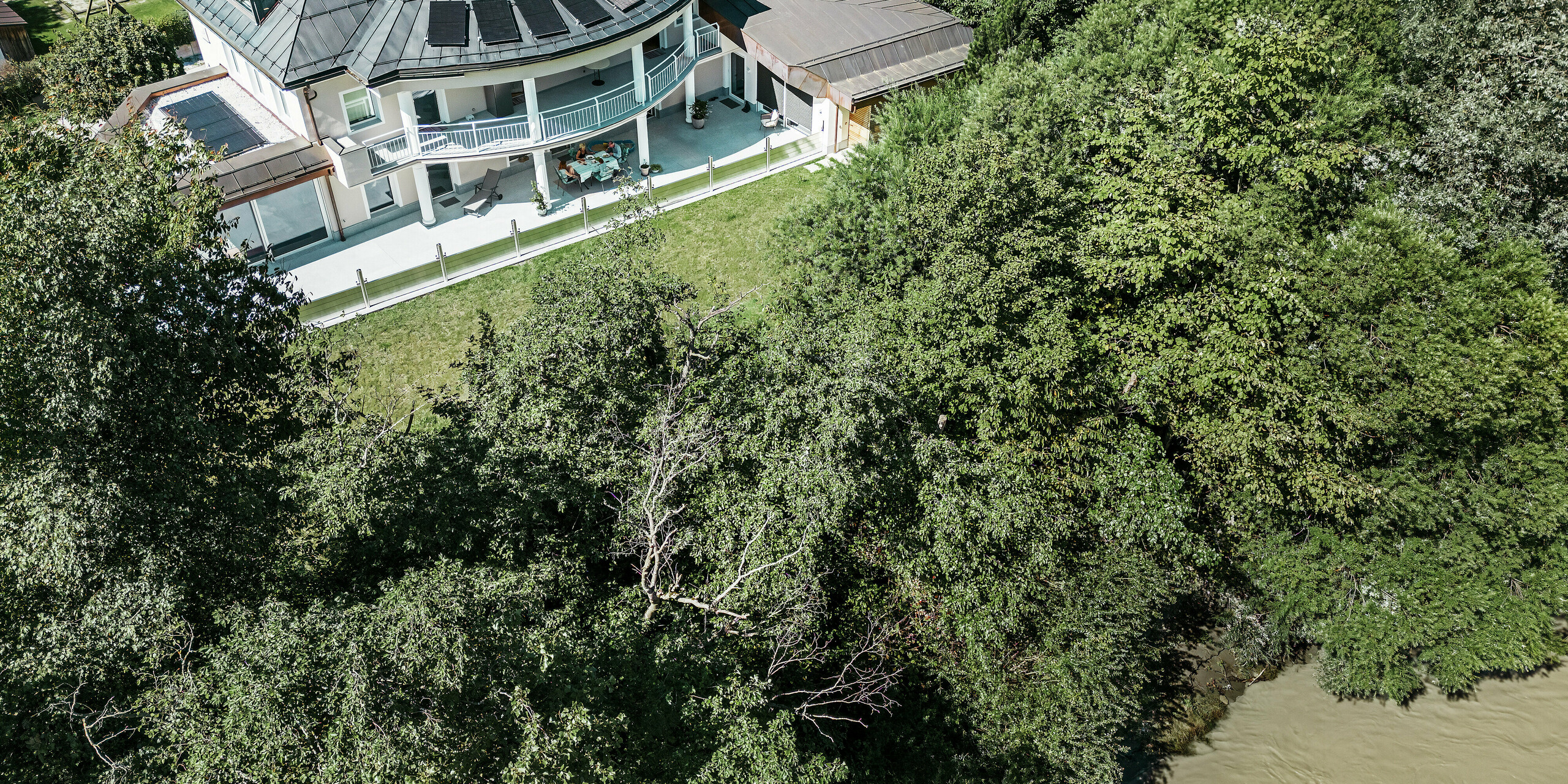 Luftaufnahme eines modernen Einfamilienhauses in Schwaz, Tirol, das direkt an einem Fluss liegt. Gut zu erkennen ist das entlang der Terrasse montierte PREFA Hochwasserschutzsystem, das mit Aluminium Dammbalken eine sichere Barriere zum angrenzenden Gewässer bildet. Die erhöhte Perspektive zeigt die unmittelbare Nähe des Hauses zum Gewässer und unterstreicht die Relevanz eines effektiven Landschafts- und Objektschutzes.