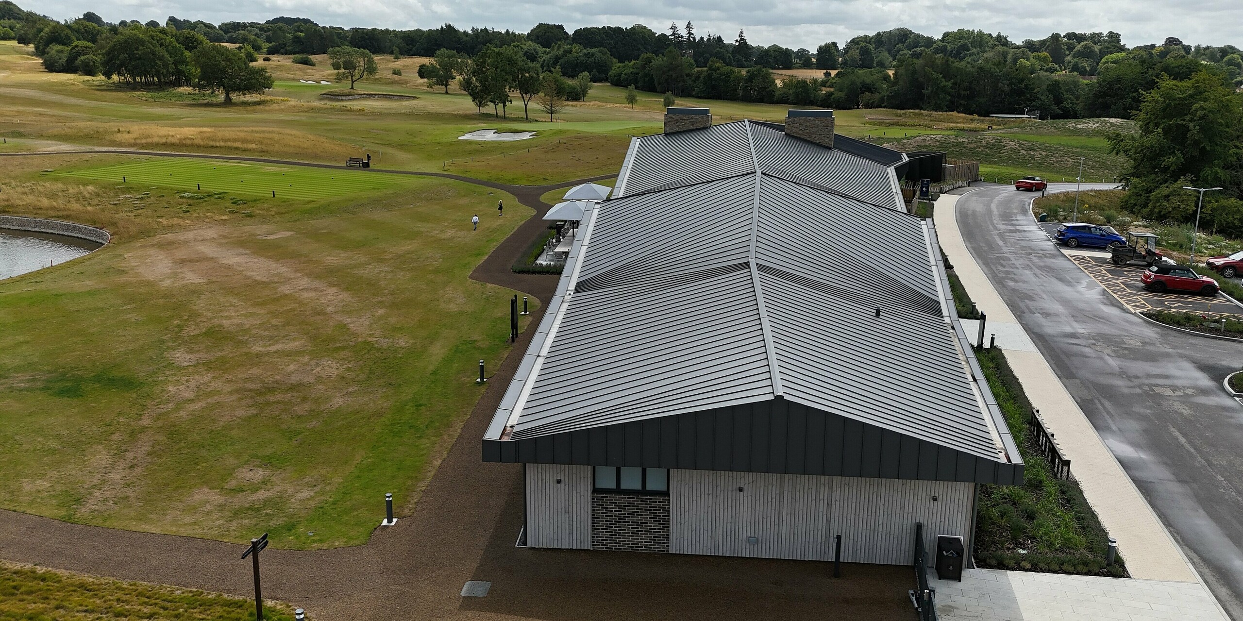 The elegant PREFALZ standing seam roof of the Basingstoke Golf Club in Hampshire attracts attention with its curved shape. The roof in P.10 dark grey creates striking accents that blend harmoniously into the expansive golf course landscape. In the foreground, you can see the gable end of the building and the well-maintained grounds of the golf course, which blend perfectly with the modern architecture of the clubhouse.