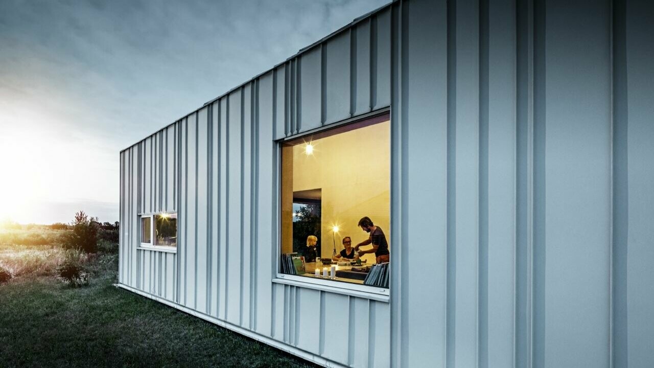Angled standing seam façade with PREFALZ in different panel widths in P.10 Prefa white photographed at dusk. Across the dining room, you can make out a family having dinner. 