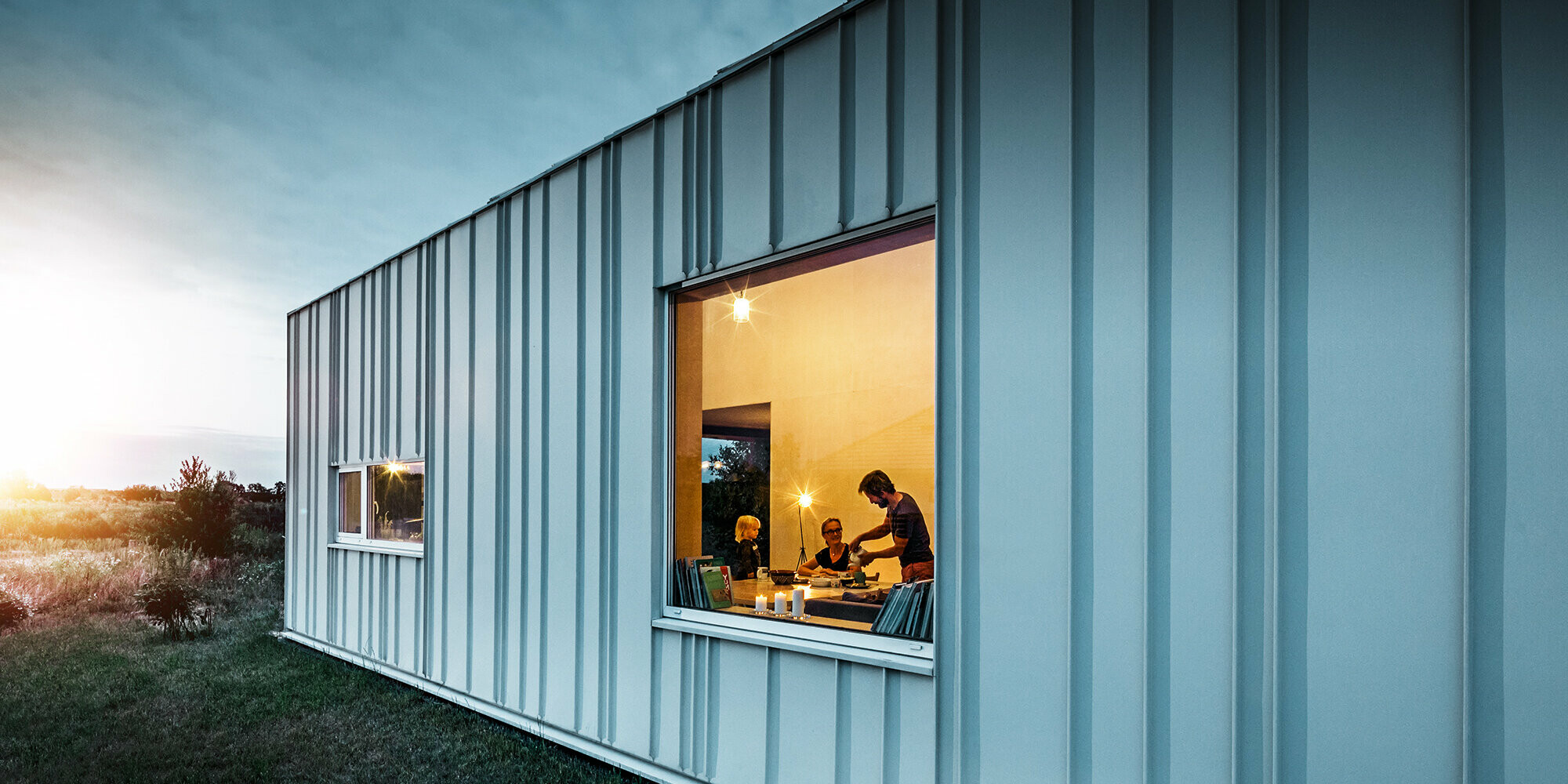 Angled standing seam façade with PREFALZ in different panel widths in P.10 Prefa white photographed at dusk. Across the dining room, you can make out a family having dinner. 