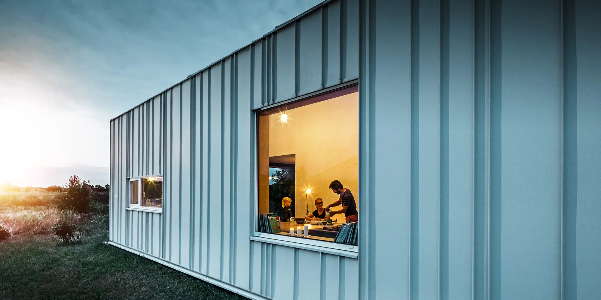 Angled standing seam façade with PREFALZ in different panel widths in P.10 Prefa white photographed at dusk. Across the dining room, you can make out a family having dinner. 